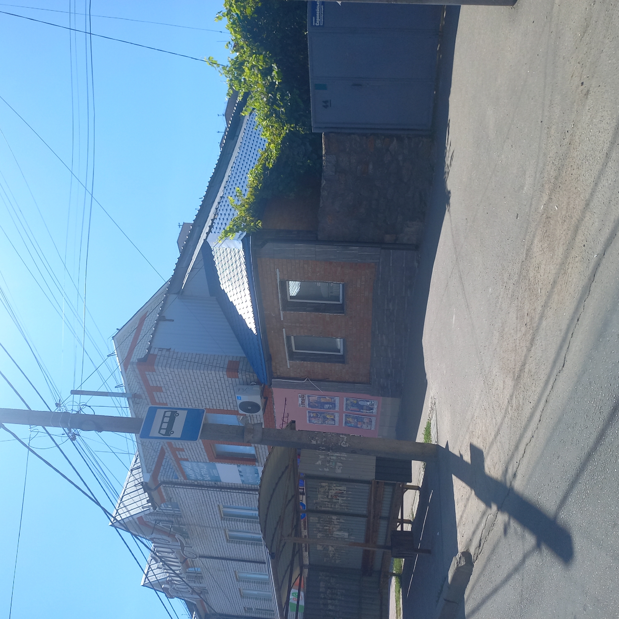 The image depicts a street scene in Kropyvnytskyi, Ukraine, on a clear, sunny day. A bus stop shelter with a corrugated metal roof and walls is visible on the left side of the frame. A pole supporting the shelter stands nearby, casting a long shadow across the asphalt road. Behind the bus stop, a multi-story building with a white brick facade and orange trim is present. A bus stop sign is affixed to a utility pole in front of this building. Further to the right, another building features a darker brick facade and a blue tiled roof. A gray metal gate with the number "44" is situated to the far right, and a street sign that reads "Серепейська" is attached to the gate. Lush green foliage encroaches on the buildings and fences from the right. The road surface is asphalt with some visible cracks and debris. The overall impression is of a typical urban environment in a temperate climate.