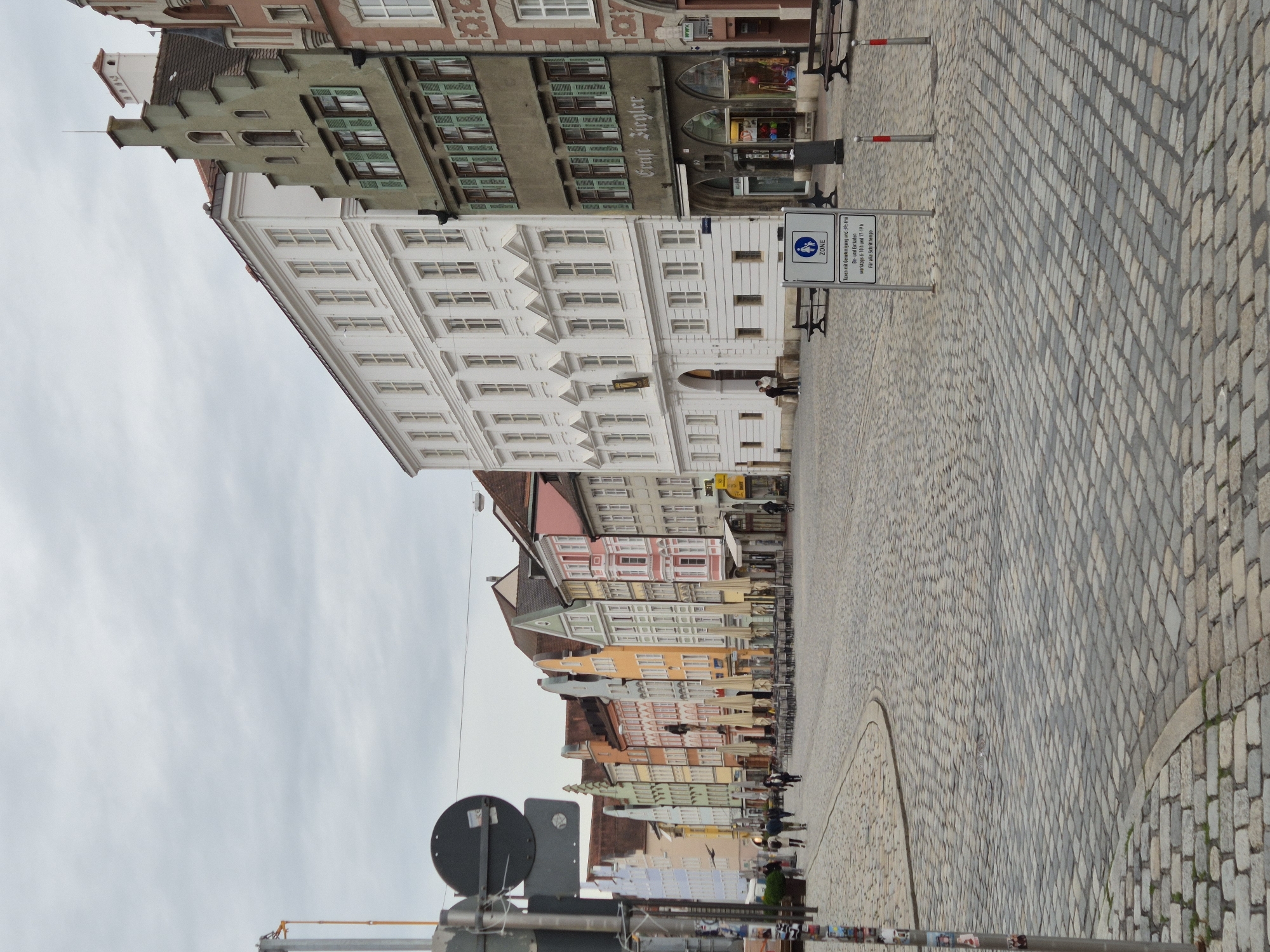 The image depicts a cobblestone street in Landshut, Germany, lined with a row of colorful buildings. The architecture is traditional European, with varied building heights and facades painted in pastel shades of pink, orange, yellow, green, and white. Some buildings feature elaborate window frames and decorative elements. Several cafes or shops have outdoor seating areas with closed umbrellas, suggesting it may be a cool or less busy time of day.
In the foreground, a metal pole with traffic signs is visible on the left. A white sign with a blue border and symbols indicating a pedestrian zone is positioned in the middle right of the image, with German text below. Benches are placed along the street. A few people are walking in the distance, and a cyclist is seen on the right side of the street. The sky is overcast, contributing to the subdued lighting of the scene. The street itself is paved with an intricate pattern of cobblestones, showing wear and variation in color. The overall impression is of a historic city center with a lively but not crowded atmosphere.