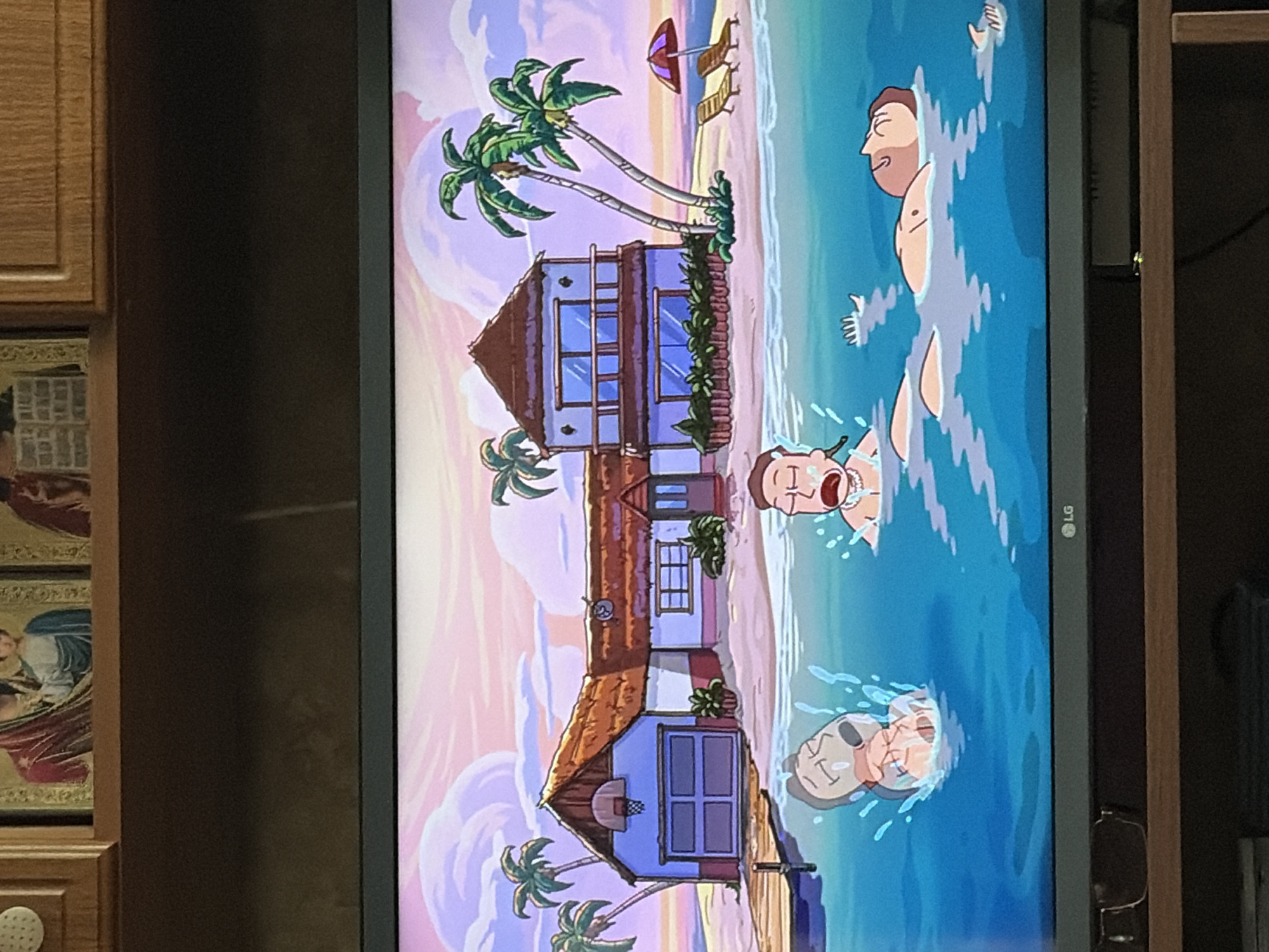 A TV screen displays a cartoon scene of a tropical island with a house and palm trees under a pink and purple sky. Three cartoon men are in the water surrounding the island. One man is submerged with only his head and arms visible, crying. Another man is partially submerged, also crying. A third man is floating on his back with his eyes closed. The house has a thatched roof, a basketball hoop above a garage, and a wooden dock extending into the water. Two lounge chairs and an umbrella are on the sandy beach.