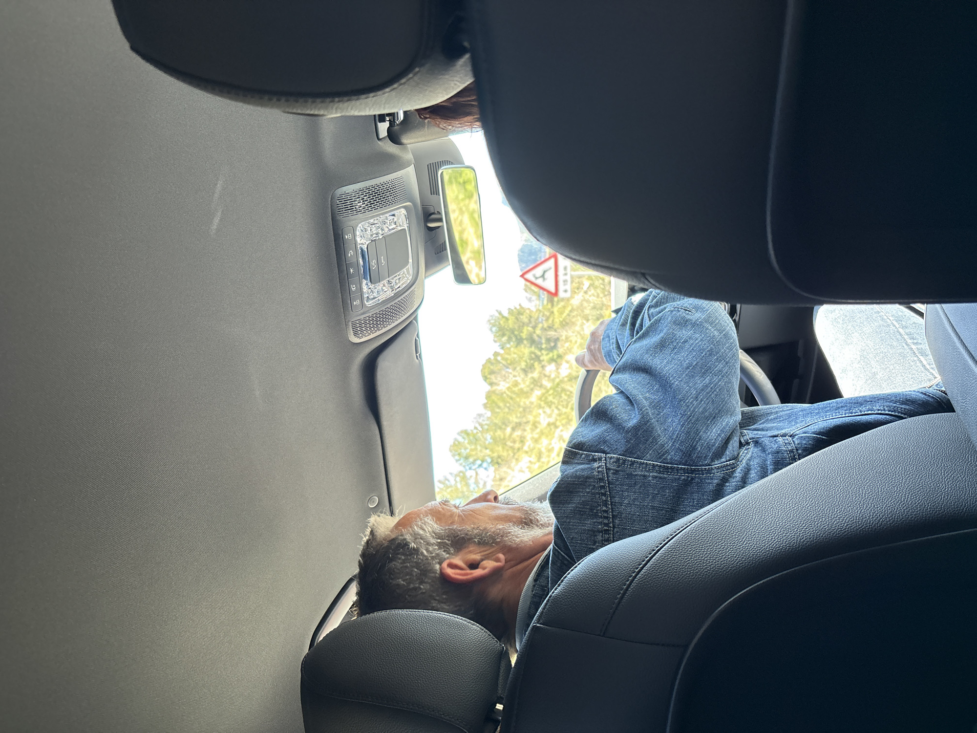 The image depicts the interior of a vehicle, likely a car or van, with a man reclined in the driver's seat. The man, who appears to be middle-aged, has graying hair and is wearing a denim shirt. He is leaning back in the seat, with his head resting on the headrest. His hands are visible on the steering wheel.
The vehicle's interior features a gray headliner and black leather seats. A rearview mirror and a panel with illuminated buttons are visible near the top of the frame.
Outside the vehicle, a glimpse of the surroundings is visible through the windshield, revealing trees and a road sign indicating a traffic island and merge, indicating that the vehicle is in transit.
The road sign visible through the windshield of the car has the letters “CV-750” written on it, indicating the car is on the CV-750 road in Benissa, Spain.