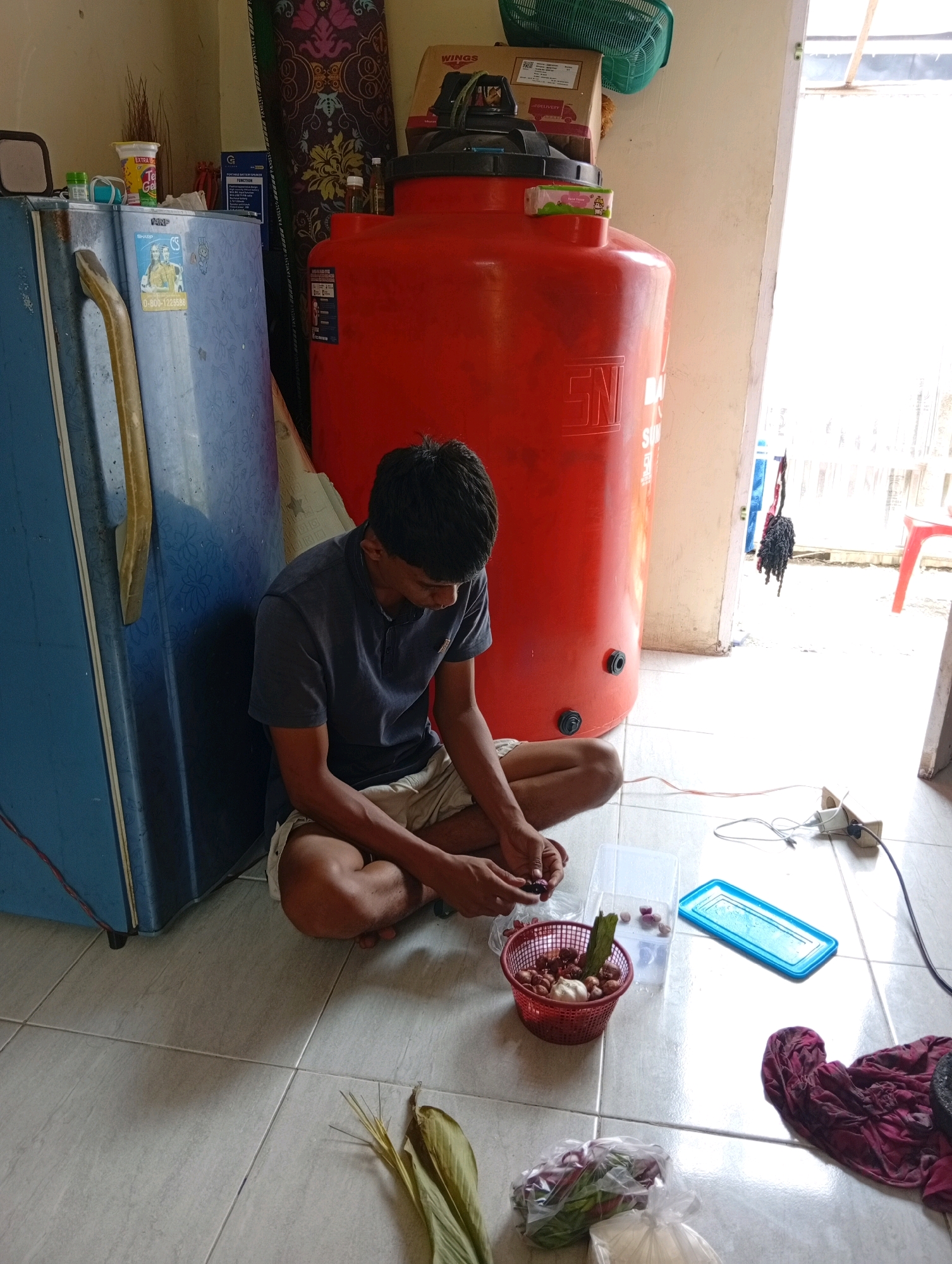 In Kualu, Indonesia, a young man sits on a tiled floor next to a large red water tank and a blue refrigerator. He is wearing a dark t-shirt and shorts, and he is engaged in preparing food, with a basket of shallots and a bulb of garlic in front of him. A knife is in his hand, and he appears to be peeling or chopping ingredients. Nearby, there are plastic containers, a blue rectangular object, and a pile of dark red cloth. Wires and an electrical outlet are visible on the floor. The environment suggests a domestic setting, possibly a kitchen or an outdoor area adjacent to a building, with a doorway in the background leading to a brighter outdoor space.