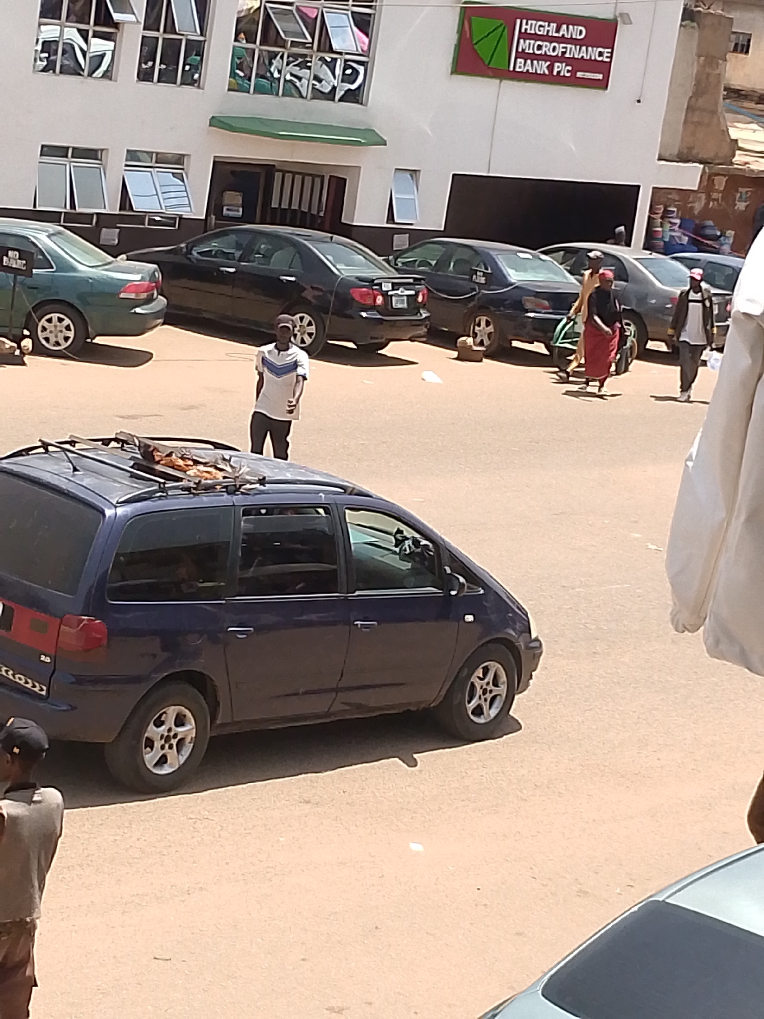 This image depicts a street scene in Bukuru, Nigeria, outside a Highland Microfinance Bank Plc. A blue minivan is parked in the foreground, its roof rack holding a wooden structure with what appears to be food items. A man in a white and blue striped shirt and dark pants stands near the minivan, looking towards the camera. In the background, several cars are parked on the street, including a dark sedan and a green sedan. People are seen walking on the sidewalk and street. The building behind them is white with multiple windows and signage for the bank. The ground is a sandy-colored surface, likely a road or parking area.