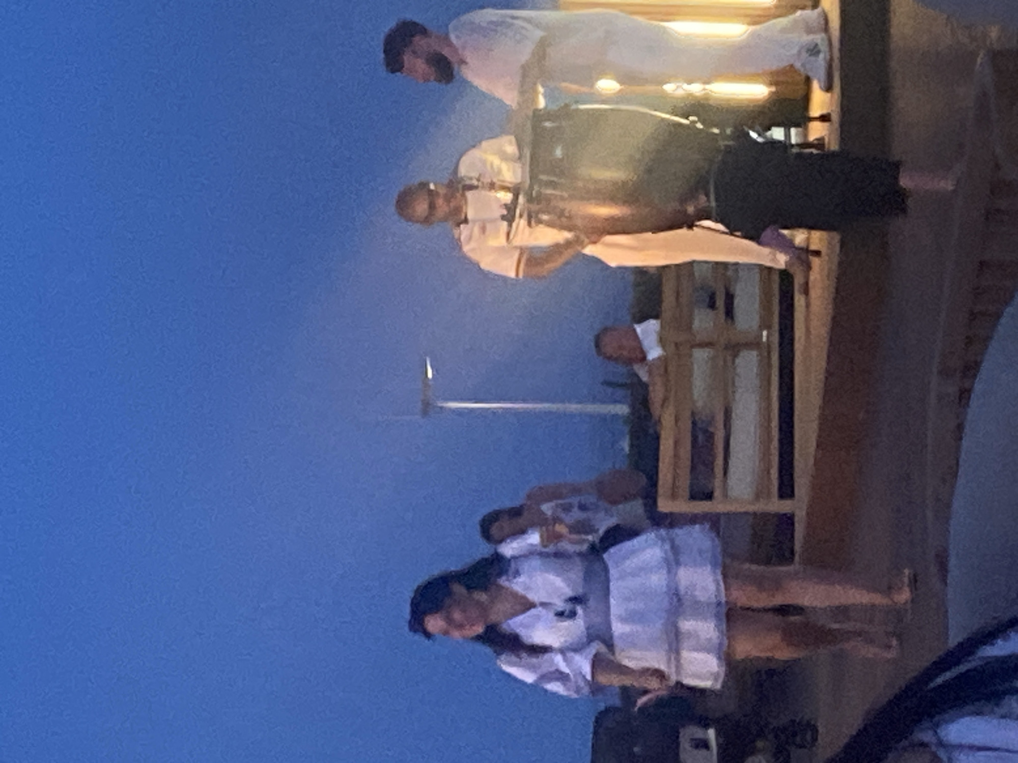 The image depicts an outdoor evening scene with a clear blue sky. In the foreground, a woman in a white tiered dress walks on a paved surface. Behind her, a woman in a patterned top holds a drink. Further back, two men dressed in white are standing near a large drum. One man appears to be playing the drum, while the other looks towards him. In the mid-ground, a person is seated on a wooden bench structure. The overall setting suggests an event or gathering in a public space, possibly a plaza or walkway in Estepona, Spain, given the lighting and attire.