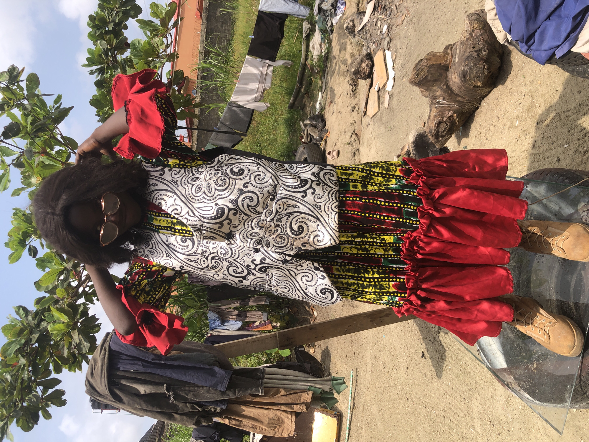 A young Black woman, appearing to be in her late teens or early twenties, stands outdoors in what appears to be a rural or informal urban setting in Ado, Nigeria. She is positioned in the foreground, facing the camera directly. She has dark, wavy hair that falls to her shoulders and is wearing rose-gold sunglasses. Her attire is a vibrant, multi-colored dress with a yellow, red, and black patterned skirt featuring a ruffled red hemline. Over the dress, she wears a white and black patterned sleeveless vest with a paisley design. Her arms are raised, and her hands are touching her hair. She is wearing tan-colored lace-up boots.
The background reveals a somewhat unkempt environment. To the left, several pieces of clothing are hanging on a wooden structure, possibly a clothesline. Behind her, a tree with large green leaves provides some shade. Further back, laundry is visible hanging on a clothesline stretched between posts. The ground is a mixture of sand and dirt, with scattered debris including pieces of wood, what looks like a discarded tire, and other assorted refuse. A low wall and vegetation are also visible in the distance. The sky is partly cloudy. The overall scene suggests a lived-in, everyday environment rather than a pristine or staged location.