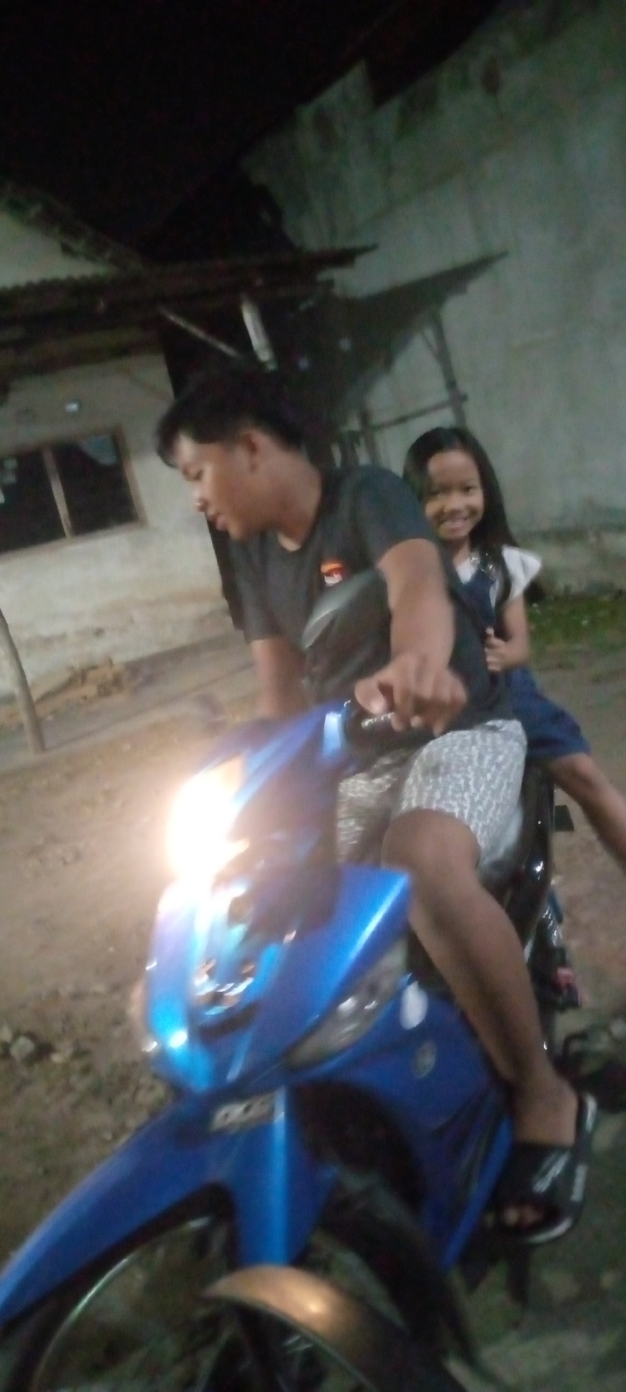 A slightly blurred, low-light, close-up shot captures a man and a young girl riding a blue motorcycle in what appears to be an outdoor, possibly rural setting. The man, in a dark t-shirt, is in the driver's seat, looking down and to the left, with his right hand on the handlebars. The girl, wearing a white top and a dark blue overall dress, is seated behind him, smiling at the camera. Her left arm is around the man's waist. The motorcycle's headlight is illuminated, casting a bright glare. In the background, a weathered building with a window is visible, along with some vegetation and what looks like a rustic structure overhead. The ground appears to be dirt or gravel. The overall impression is a casual, perhaps evening, ride. The specific location in Sumberwono, Indonesia, is not explicitly depicted but is assumed to be a general area with a mix of rural elements and basic structures.
