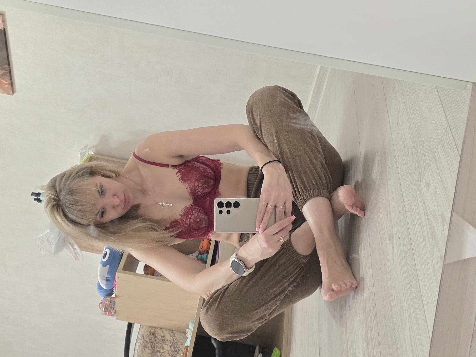 A woman with blonde hair and bangs takes a mirror selfie in an indoor setting, likely a home in Chaikovsky, Russia. She is seated on a light wooden floor with her legs bent, wearing a maroon lace bra and brown, textured sweatpants. She holds a smartphone, identifiable by its camera module (resembling a Samsung Galaxy S23 Ultra), angled towards the mirror. A necklace with a cross pendant hangs around her neck, a light-colored smartwatch is on her left wrist, and she wears several rings on her right hand, which also has light-painted fingernails. Her gaze is directed towards the phone. The background, reflected in the mirror, features white walls and a light wooden shelf or cabinet holding a blue and white plush toy (possibly a shark), a container of colorful bands, a white plastic bag, and a framed picture. The room appears well-lit, but the specific time of day is not indicated. No visible text is present in the image.