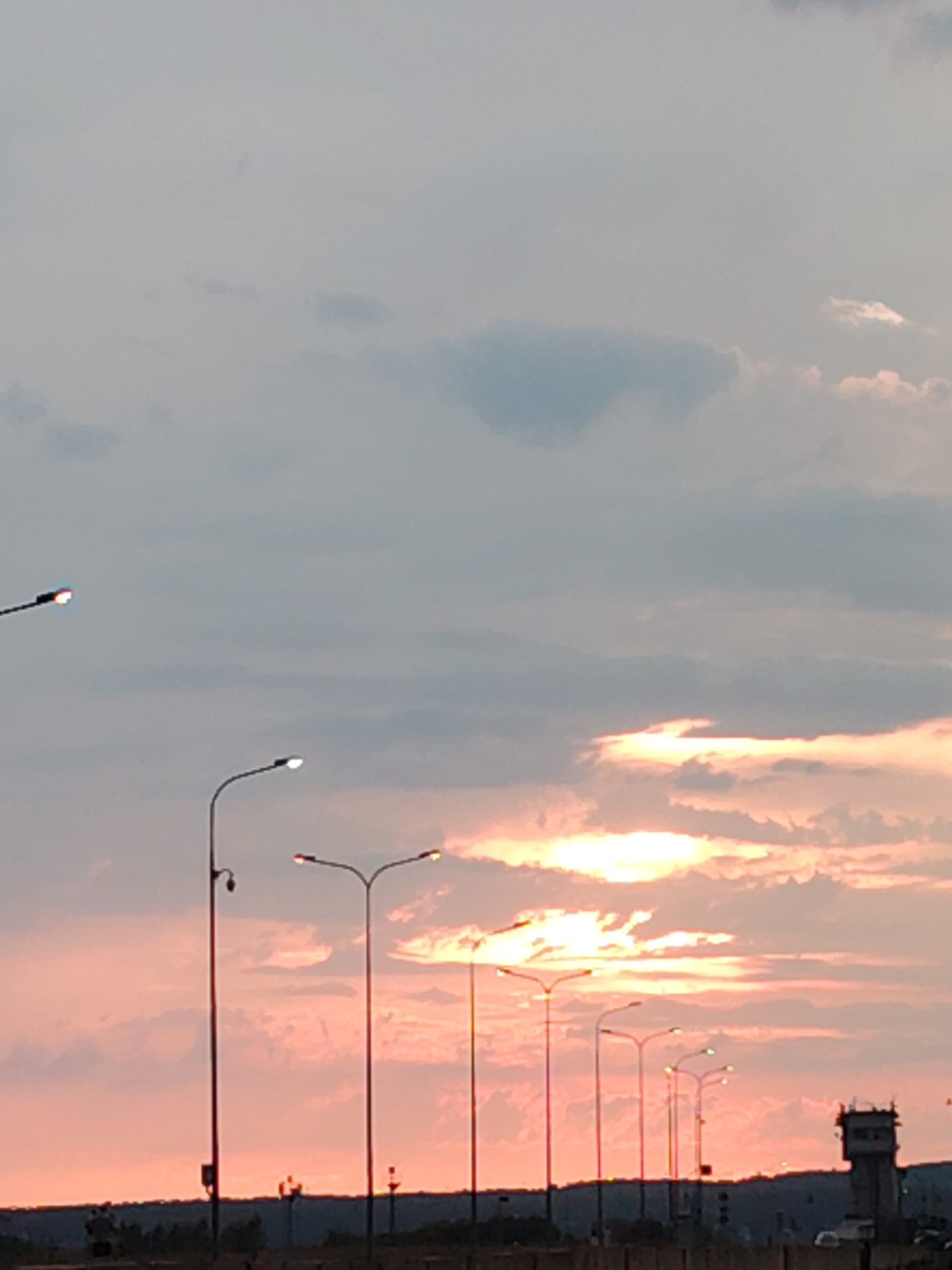 The image captures a sunset scene with a line of streetlights along a raised structure, possibly a bridge or highway. The sky is filled with soft clouds tinged with the warm hues of the setting sun, ranging from pale pink to vibrant orange and yellow where the sun's rays are breaking through. The light creates a dramatic and serene atmosphere.
Several streetlights are visible, some illuminated with a bright white light, others with a reddish glow, suggesting a transition from day to night. The streetlights are tall and slender, with multiple lamps on each pole. In the background, a dark silhouette of trees and a structure resembling a control tower or small building can be seen against the colorful sky. The overall setting appears to be an urban or semi-urban environment, with the elevated road suggesting a transportation infrastructure.
There are no visible people in the foreground or middle ground, though there might be some very small figures in the distant background. The time of day is clearly sunset, indicated by the warm, low-angle sunlight and the colors in the sky. The weather appears calm, with scattered clouds. The location cues, combined with the provided context of Chaikovsky, Russia, place the scene there. The image evokes a sense of calm and the beauty of natural light at the end of the day. No visible text is present in the image.