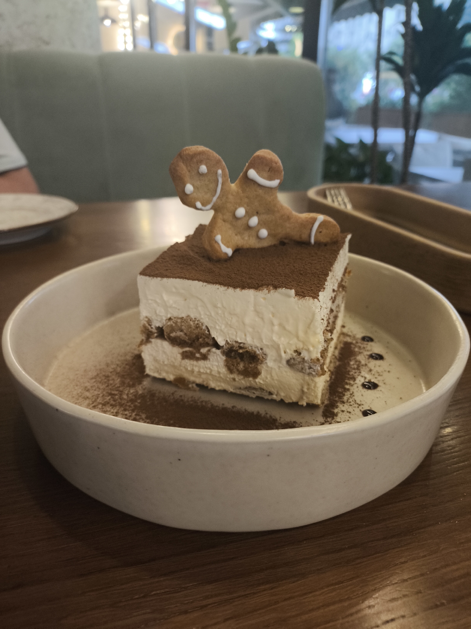 A square slice of tiramisu topped with cocoa powder and a gingerbread man cookie sits in a white bowl on a wooden table. The tiramisu has layers of coffee-soaked ladyfingers and mascarpone cream. The gingerbread man is decorated with white icing for eyes, a smile, and buttons, and is positioned with its arms and legs splayed. In the background, out of focus, is a green upholstered bench, a blurry figure in a light-colored shirt, a large window with greenery outside, and what appears to be the interior of a cafe or restaurant. The overall lighting suggests an indoor setting, possibly during the day. The environment appears to be a relaxed dining area. No specific location in Saint Petersburg, Russia, can be identified from the image.