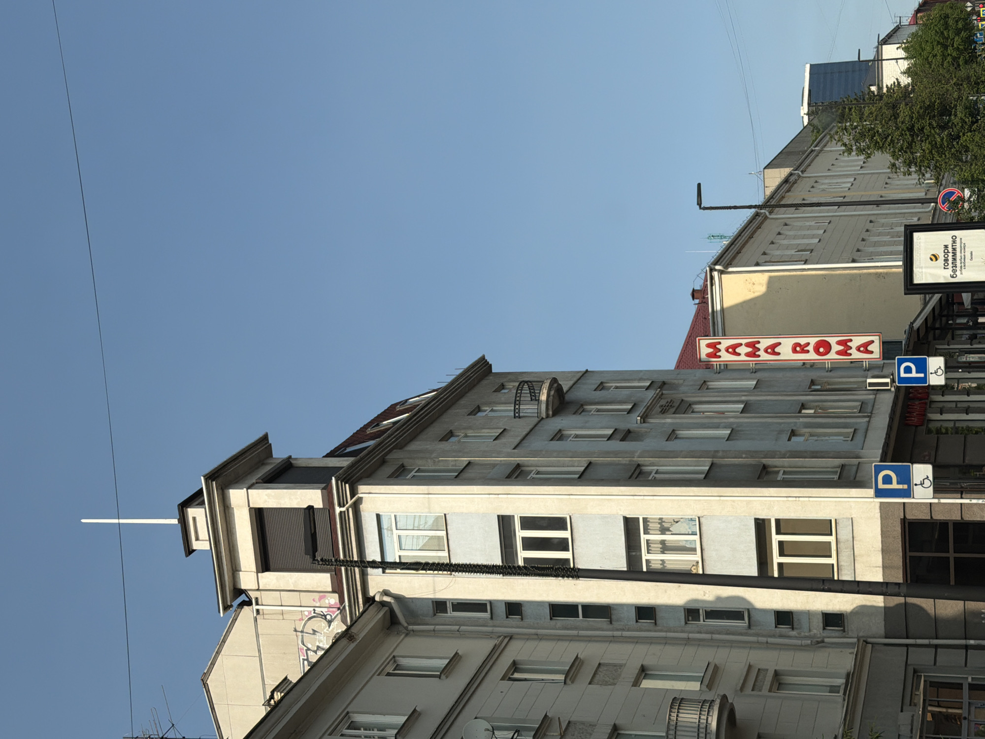 The image shows a street view in Krasnoyarsk, Russia, on a clear day with a bright blue sky. In the foreground, a tall, slender lamppost is positioned to the left of the frame, casting a shadow on a pale blue building. This building has multiple floors with numerous windows, some of which are partially covered by curtains. Above this building, to the right, is a yellow building with a prominent vertical red sign that reads "MAMA ROMA". Next to the "MAMA ROMA" sign, there is a blue and white parking sign with a wheelchair symbol. Further to the right, a modern blue glass skyscraper is visible in the distance, surrounded by trees. Another, lighter-colored building stands between the yellow "MAMA ROMA" building and the skyscraper. The architectural style of the older buildings suggests an urban setting with a mix of historical and contemporary structures. The overall scene depicts a typical street in a Russian city.