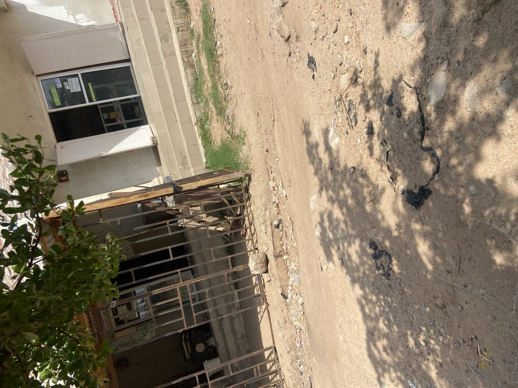 The image depicts a street scene in Jalingo, Nigeria, with a focus on the exterior of a commercial establishment. A dirt road, scattered with rocks and debris, occupies the foreground, with shadows of tree leaves dappled across it. To the right, a building with a white facade and a storefront is visible, featuring double glass doors and a few steps leading up to the entrance. A few signs are affixed to the glass doors. To the left, a metal gate bars access to an area containing what appears to be a generator and some shelves behind it, suggesting a storage or workshop space. The overall impression is one of a somewhat unkempt urban environment, characteristic of many parts of Jalingo.