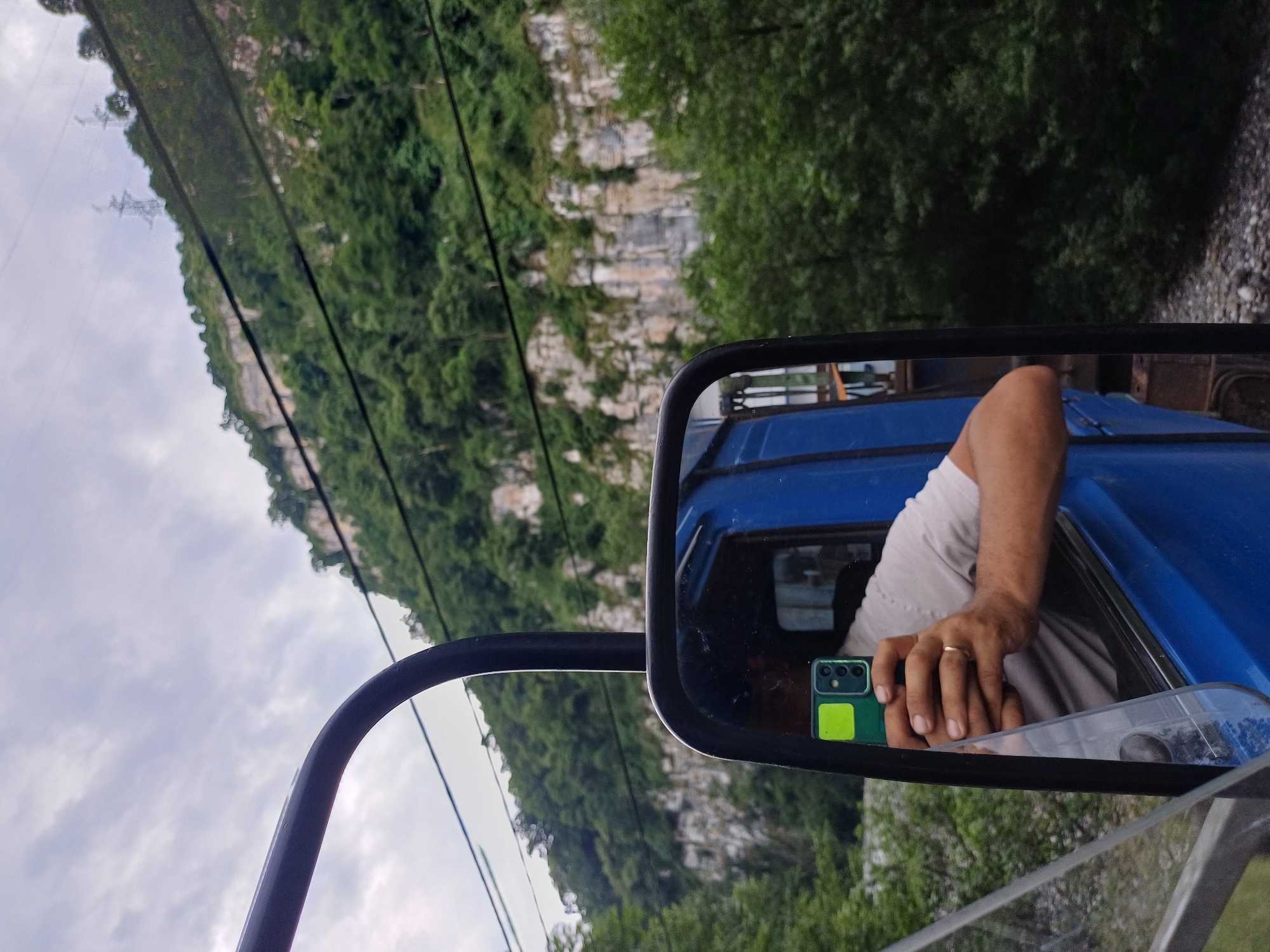 A person is holding a smartphone in their right hand, reflected in the side-view mirror of a blue vehicle. The smartphone is green with a yellow sticker on its back. The person is wearing a light-colored shirt and a ring on their left hand. The reflection in the mirror shows a mountainous landscape with lush green trees and a cloudy sky. The photo appears to be taken from inside the vehicle, looking out and then at the reflection. The scene suggests a travel or road trip in a mountainous region, possibly during the daytime given the lighting. The location context is Adlerskiy, Russia.