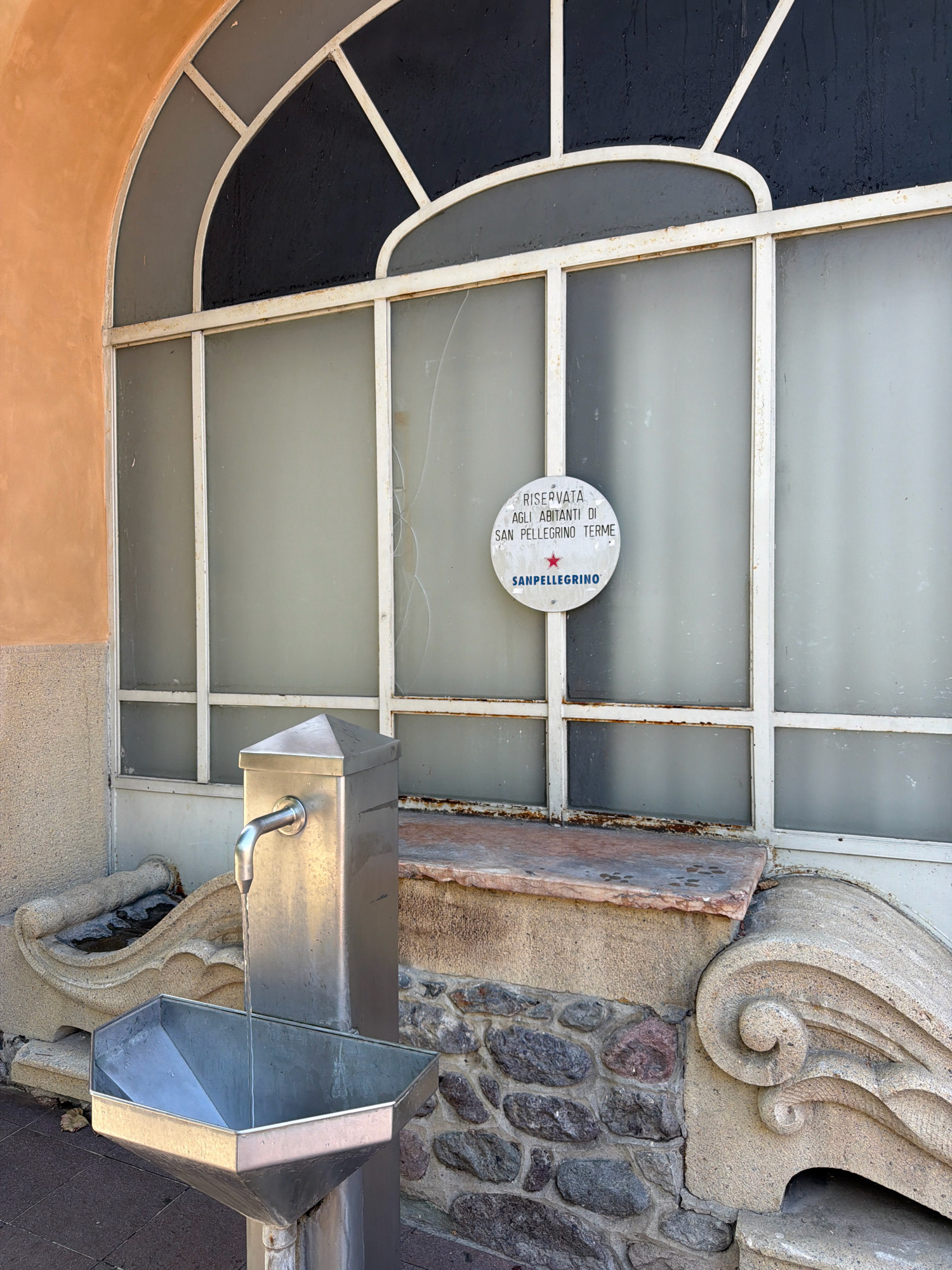The image shows a public water fountain in San Pellegrino Terme, Italy. The fountain is made of stainless steel with a spigot and a basin. Behind the fountain is a large arched window with frosted glass panes. A circular sign on the window reads "RISERVATA AGLI ABITANTI DI SAN PELLEGRINO TERME" and also displays the Sanpellegrino logo. The fountain is located in front of a building with an orange stucco facade and a stone base. The surrounding environment includes a paved walkway and decorative stone carvings.