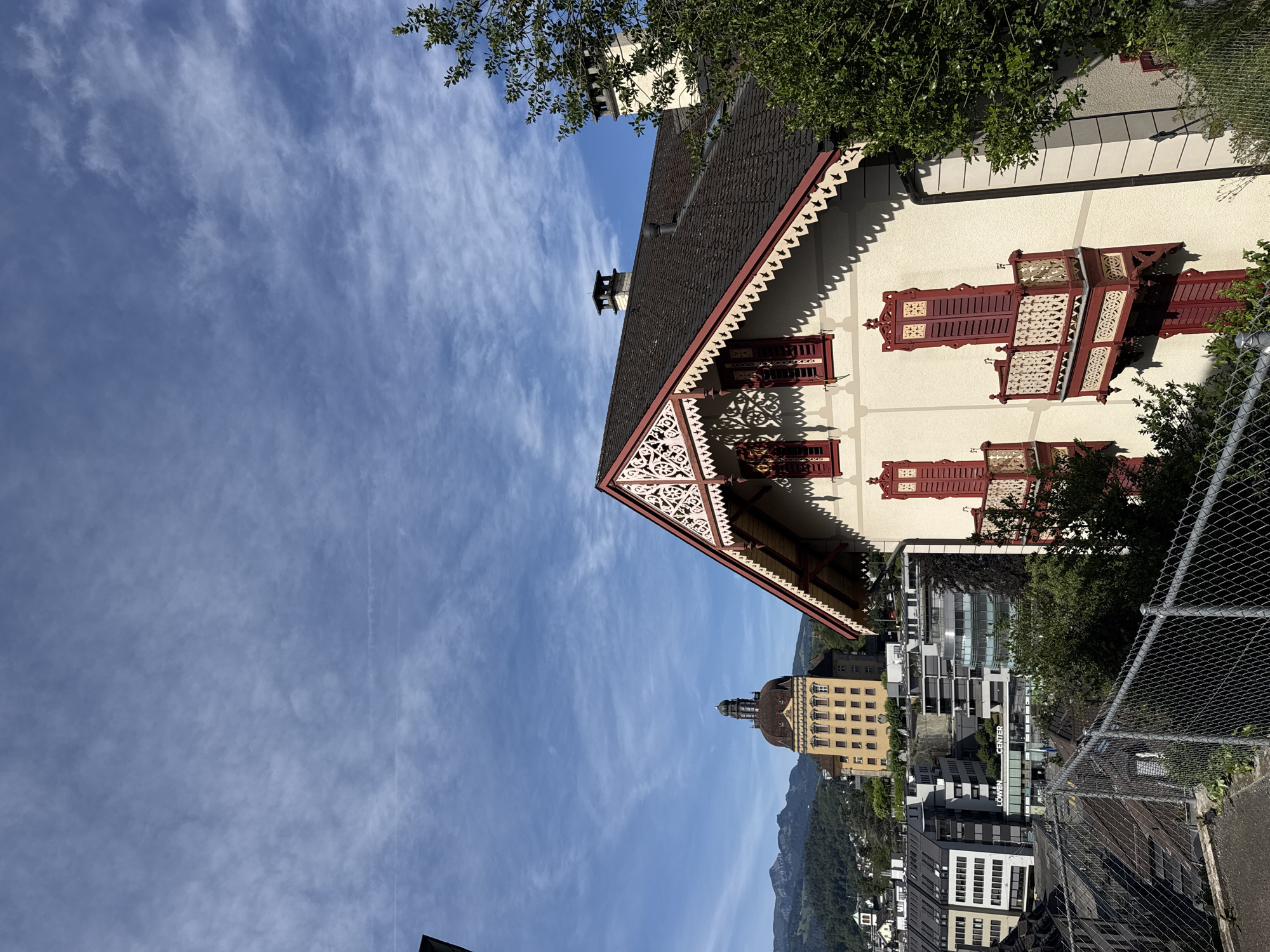 This is a high-angle, medium shot of the city of Lucerne, Switzerland, on a clear, partly cloudy day. In the foreground, a two-story building with a red-shingled roof and white trim features ornate decorative gables with intricate lattice work. Red-shuttered windows and small balconies with decorative railings are visible on the facade. To the left and behind this building, a chain-link fence runs across the bottom of the frame. In the middle ground, the "Löwen Center" building, a modern structure with glass and concrete, is visible, along with other contemporary buildings. Further in the background, a large, yellow building with a domed roof and tower stands prominently on a hillside, with lush green trees and mountains in the distance. The sky is a bright blue with scattered white clouds.