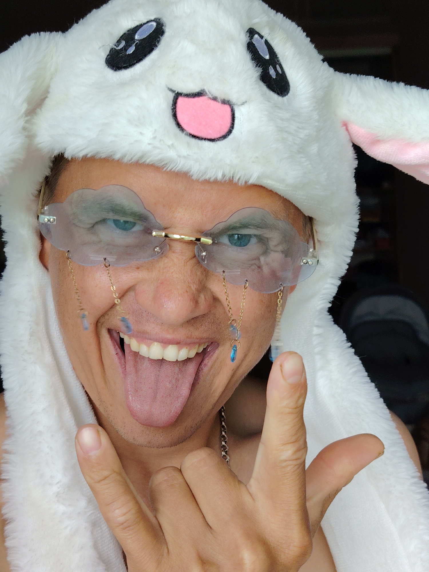 A person wearing a white plush animal hat with embroidered eyes and a pink mouth is sticking their tongue out and making a rock on hand gesture. They are also wearing cloud-shaped glasses with chains and blue gem-like charms. The image is a close-up portrait of the person's face and upper torso. There is no discernible background detail that would indicate a specific location in Chaikovsky, Russia.