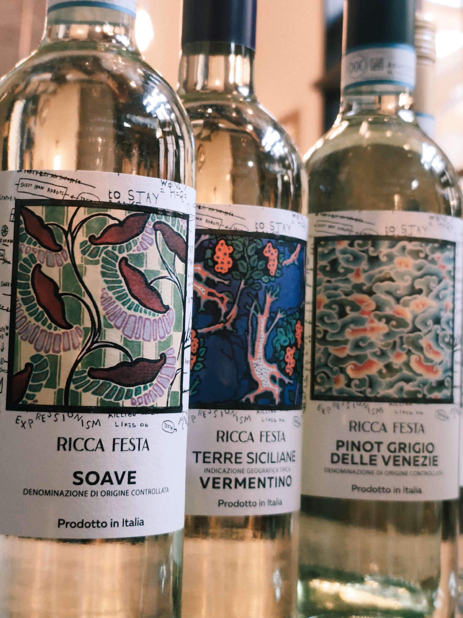 This media file is a close-up, eye-level shot of three bottles of white wine, arranged side-by-side and slightly out of focus. The bottles are made of clear glass and are filled with pale yellow wine. Each bottle features a distinctive label with an artistic design and text.
The labels share a common theme, with the words "RICCA FESTA" prominently displayed. Below this, the specific wine varietal is listed: "SOAVE" on the left, "TERRE SICILIANE VERMENTINO" in the middle, and "PINOT GRIGIO DELLE VENEZIE" on the right. All labels also include the phrase "DENOMINAZIONE DI ORIGINE CONTROLLATA" or "INDICAZIONE GEOGRAFICA TIPICA" and "Prodotto in Italia," indicating their Italian origin.
The artwork on each label is stylized and appears to be hand-drawn. The left bottle's label features a pattern of dark red, leaf-like shapes against a green and pale purple background with floral elements. The middle bottle's label depicts a whimsical, tree-like figure with orange berries against a deep blue background. The right bottle's label displays an abstract, swirling pattern in shades of blue, green, and pink, resembling clouds or water. Small, handwritten annotations and drawings are visible around the main artwork on the labels, adding a personal and artistic touch.
The lighting suggests an indoor setting, possibly a wine shop or a tasting room, with soft light illuminating the bottles and creating reflections on the glass. The background is blurred, further emphasizing the wine bottles as the primary subjects. No people are visible in the image. The overall mood is sophisticated and artistic, highlighting the unique branding of the wines.