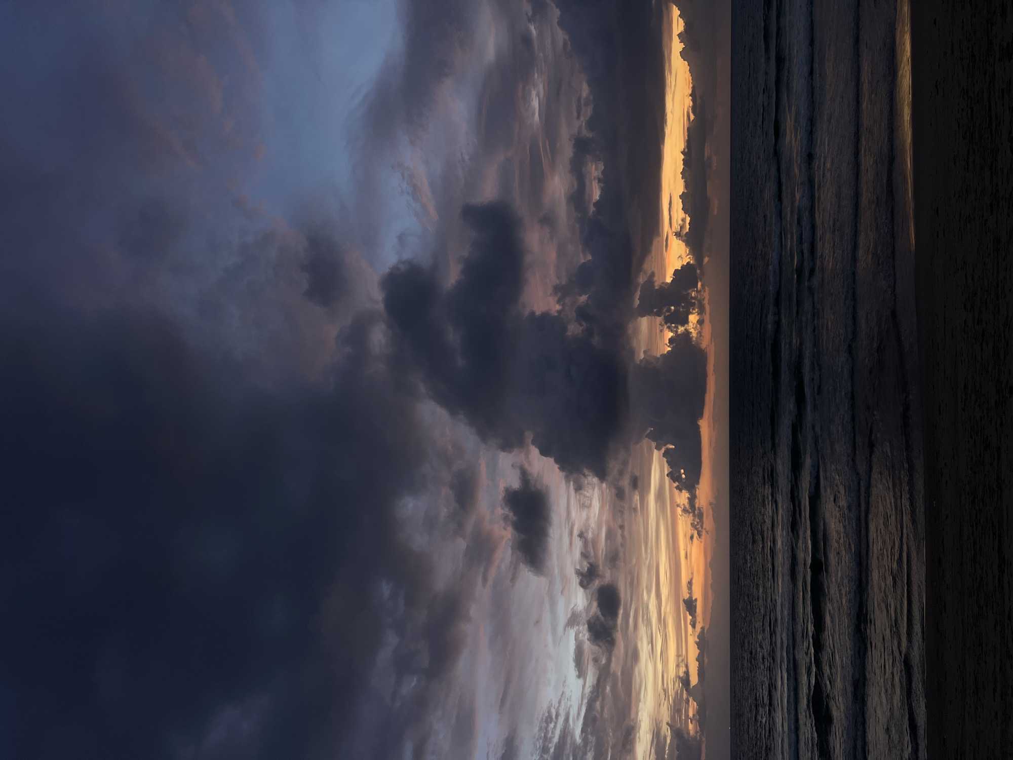 This is a photograph taken at dusk on the beach in Choeng Thale, Thailand. The sky is filled with dramatic clouds in various shades of dark blue, purple, and orange, illuminated by the setting sun. The ocean is dark with visible waves rolling towards the shore. The foreground is a dark, sandy beach. There are no people or man-made objects visible in the frame. The overall atmosphere is moody and serene.
