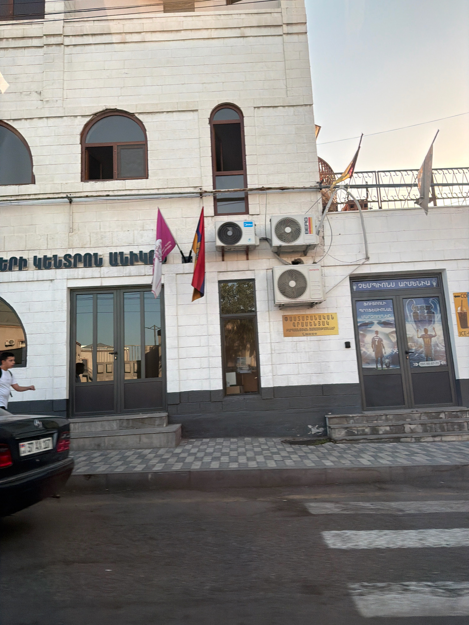 The image captures a street-level view of a building in Yerevan, Armenia, likely situated on a commercial street given the presence of storefronts and signage. The building is constructed with white brick and features multiple arched windows. Two main entrances are visible, one with double glass doors and another to the right, also with double glass doors, which appear to be the entrance to a shop or establishment. Above the left entrance, text in Armenian is displayed. On the facade, several air conditioning units are mounted. To the right of the entrance, a golden plaque with Armenian text is affixed to the wall. The right entrance features posters of soccer players and a trophy, suggesting a sports-related business. Three flags are displayed on poles above the building, two of which are visible in their entirety. A pedestrian crossing is marked on the asphalt road in the foreground. A black car is parked on the left side of the image, with its rear visible, displaying a license plate. A person in a white t-shirt and dark pants is walking near the left entrance. The sky above is clear and appears to be late afternoon or early evening, with a warm glow.