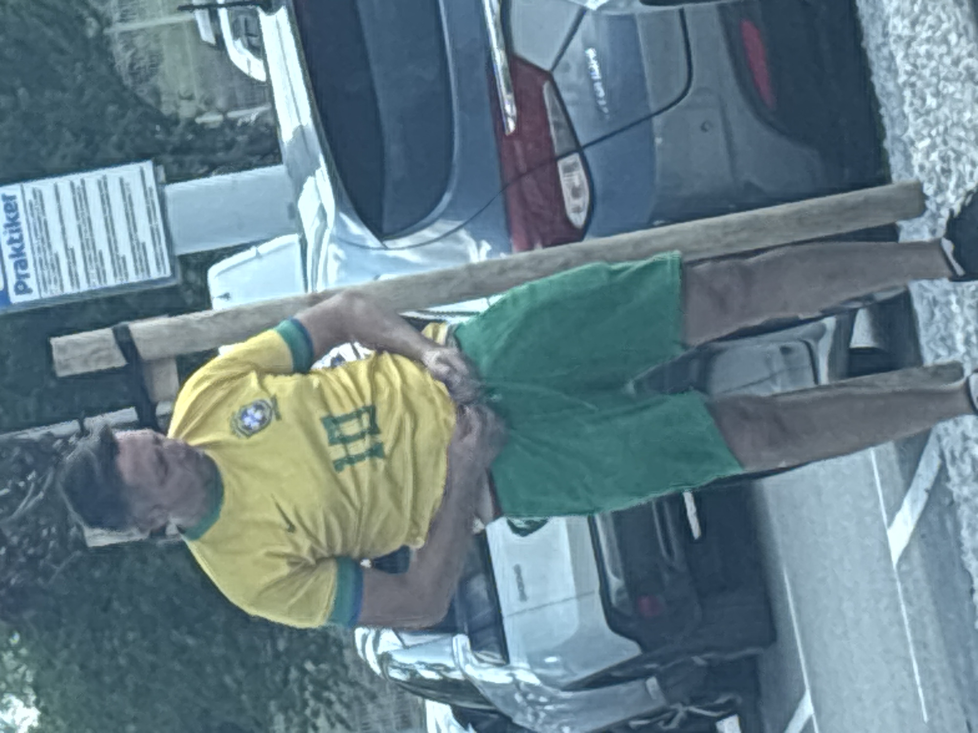 An outdoor scene in Sofia, Bulgaria, depicts a male individual, appearing middle-aged, adjusting his attire. He is positioned centrally, wearing a yellow short-sleeved t-shirt with green and blue trim, featuring a Nike swoosh logo on the right chest and the Brazilian Football Confederation (CBF) crest on the left, along with the number "10" on the front. He is also wearing green shorts, which he holds at the waistband with both hands as if adjusting them. He wears dark shoes with white socks.
In the background, multiple vehicles are visible. A light-colored SUV, consistent with a Peugeot 3008, is parked to the left and behind the man. To his right and further back, a darker sedan, possibly a Hyundai Elantra, is parked. Above the sedan, a white sign with a blue top header displaying "Praktiker" is visible, with multiple lines of text underneath, primarily in German, likely detailing parking regulations. A light-colored wooden pole stands vertically to the right of the man. The ground consists of asphalt with white markings, transitioning to a surface with light gravel or small stones in the foreground. Green foliage from trees or bushes is present in the distant background. The scene is illuminated by natural daylight.