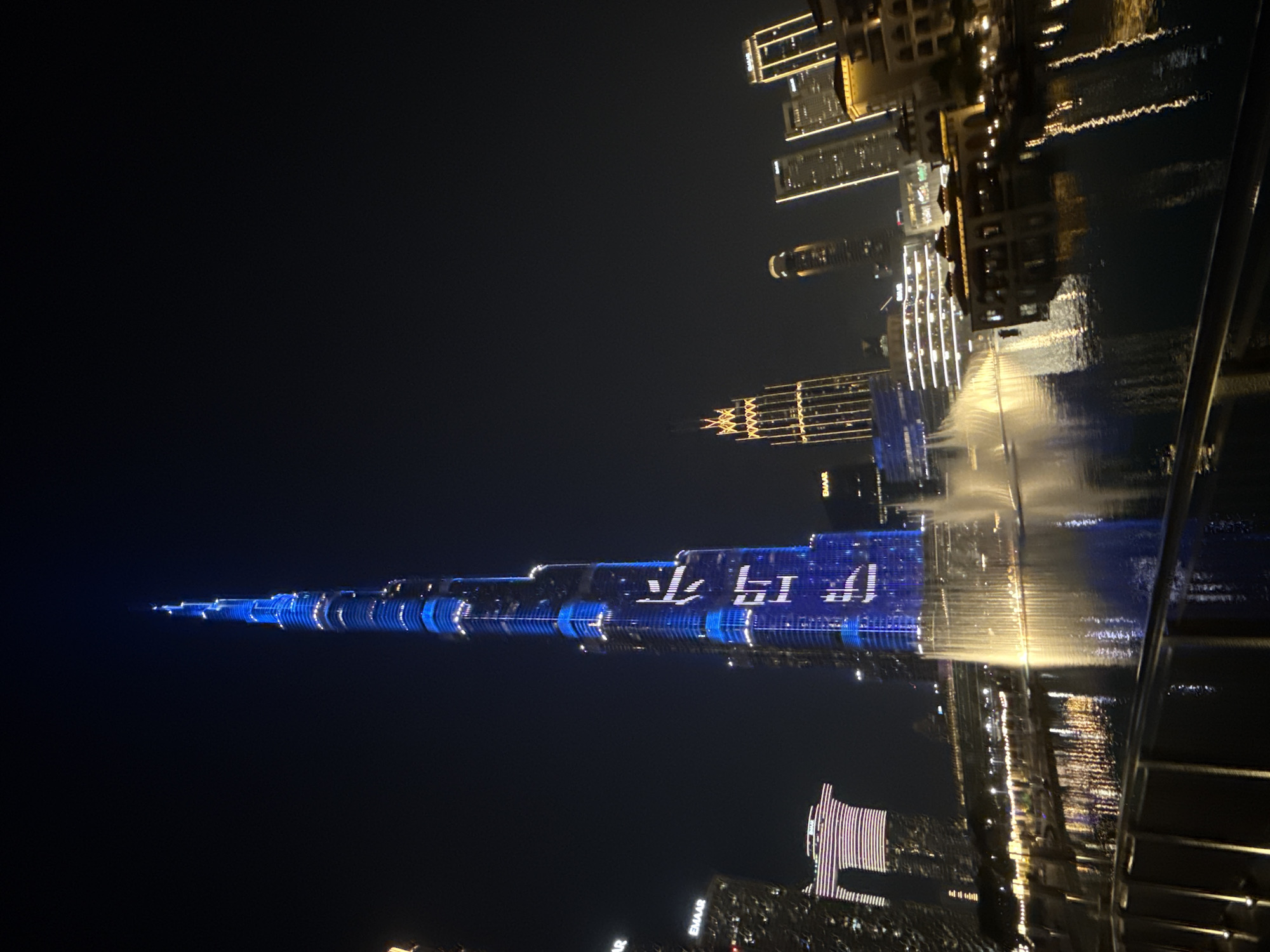 At night, the Burj Khalifa in Dubai, United Arab Emirates is illuminated with blue lights and projected with white Chinese characters. Other illuminated buildings stand in the background, with some reflected in a body of water.