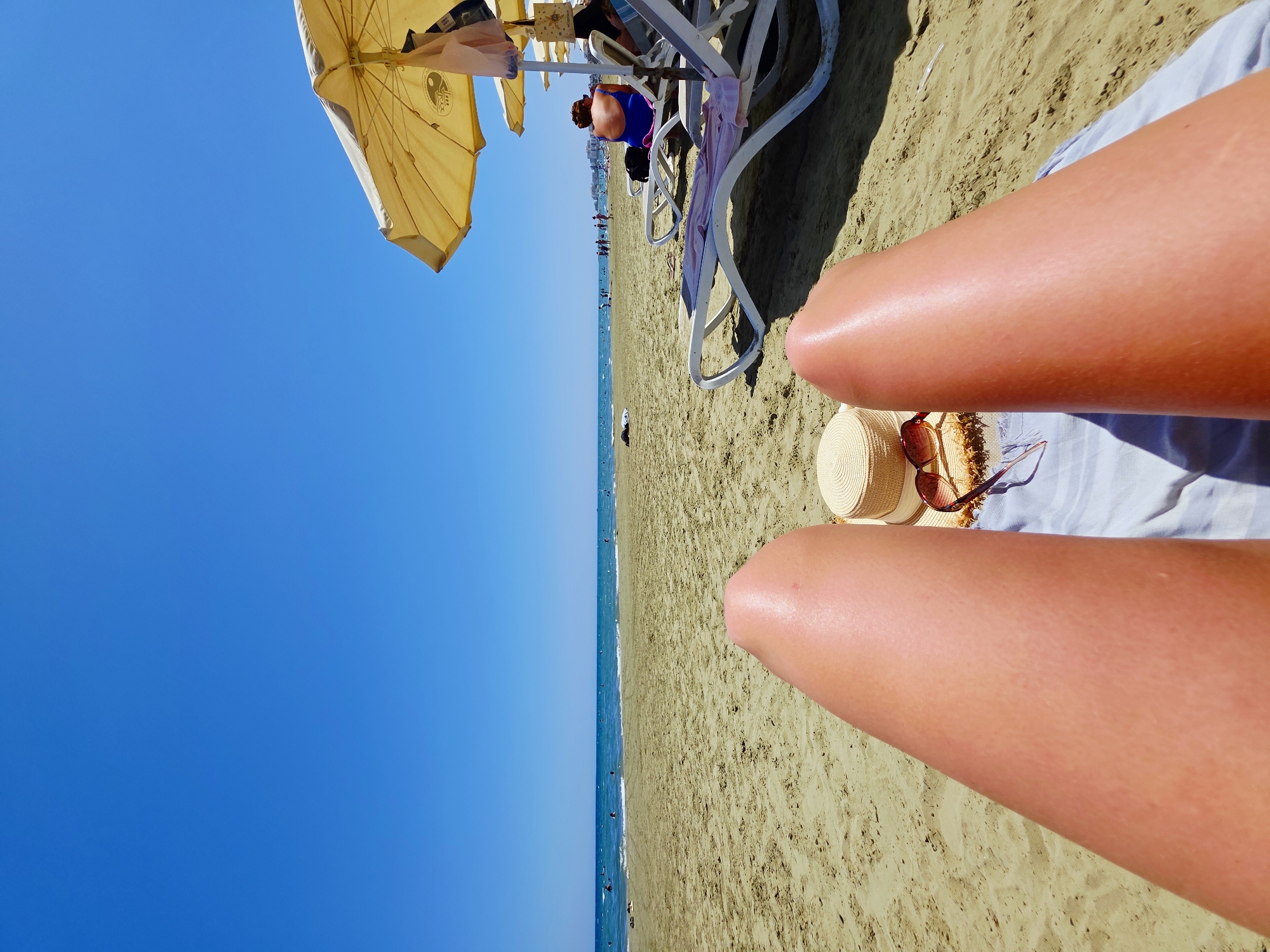 The image shows a first-person perspective of someone relaxing on a beach in Larnaca, Cyprus. In the foreground, a person's legs are visible, resting on a light blue towel. Next to the towel are a straw hat and a pair of sunglasses. The sandy beach stretches out to the sea, where several people are swimming and walking in the shallow water. In the background, beach umbrellas and lounge chairs are set up, and a few more people are visible on the beach and in the water. The sky is clear and blue, indicating a sunny day. The overall scene depicts a typical beach day with people enjoying the sun and the sea.
