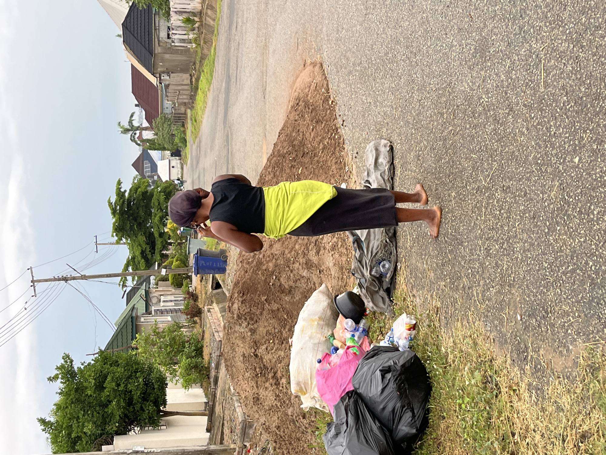The image captures a street scene in Abuja, Nigeria, during the daytime under an overcast sky. In the foreground, a person, likely female, stands barefoot on a paved road next to a mound of dirt and several piles of trash. She is wearing a dark short-sleeved top, dark trousers, a bright neon-yellow apron or wrap around her waist, and a dark head covering. Her posture suggests she is looking down or engaged in an activity related to the trash, possibly collecting or sorting waste.
To her right, a collection of waste is visible, including black trash bags, a white sack filled with items, loose plastic bottles, a black bucket, and other debris on the ground. A blue trash bin, partially visible behind her, has the text "PLOT 11SA" written on its side.
The road surface is asphalt, and beyond it, green grass and wild vegetation grow. In the background, there are several residential buildings with varied architectural styles and roof colors (dark shingle, red, green). Tall green trees and shrubs are scattered throughout the scene, and utility poles with multiple power lines stretch across the sky, connecting various parts of the neighborhood. The overall impression is one of a residential area with visible waste management challenges.