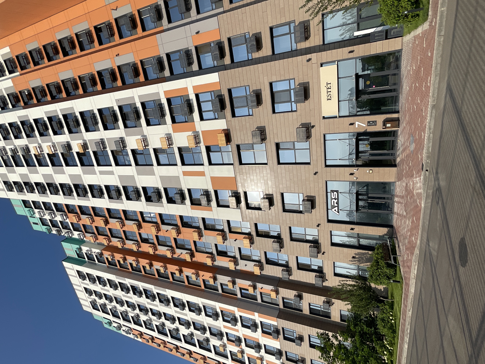 The image shows the facade of a modern, multi-story residential building with a distinct architectural style, located in Almaty, Kazakhstan. The building features a contemporary color palette with sections of white, gray, orange, and a touch of green near the top. Numerous windows and air conditioning units are visible on each floor, arranged in a regular pattern. The lower level of the building has a commercial storefront with glass doors and signage. One sign reads "ARS" in large letters, and another, a tan banner, displays "ESTÉT BEAUTY CLINIC." A building number "7" is also visible near one of the entrances. The ground in front of the building is paved with reddish-brown brickwork and includes a sidewalk and a paved road. Greenery, including trees and bushes, surrounds the base of the building and lines the sidewalk, suggesting a well-maintained urban environment. The sky above is clear and blue, indicating a sunny day. No people are present in the frame.