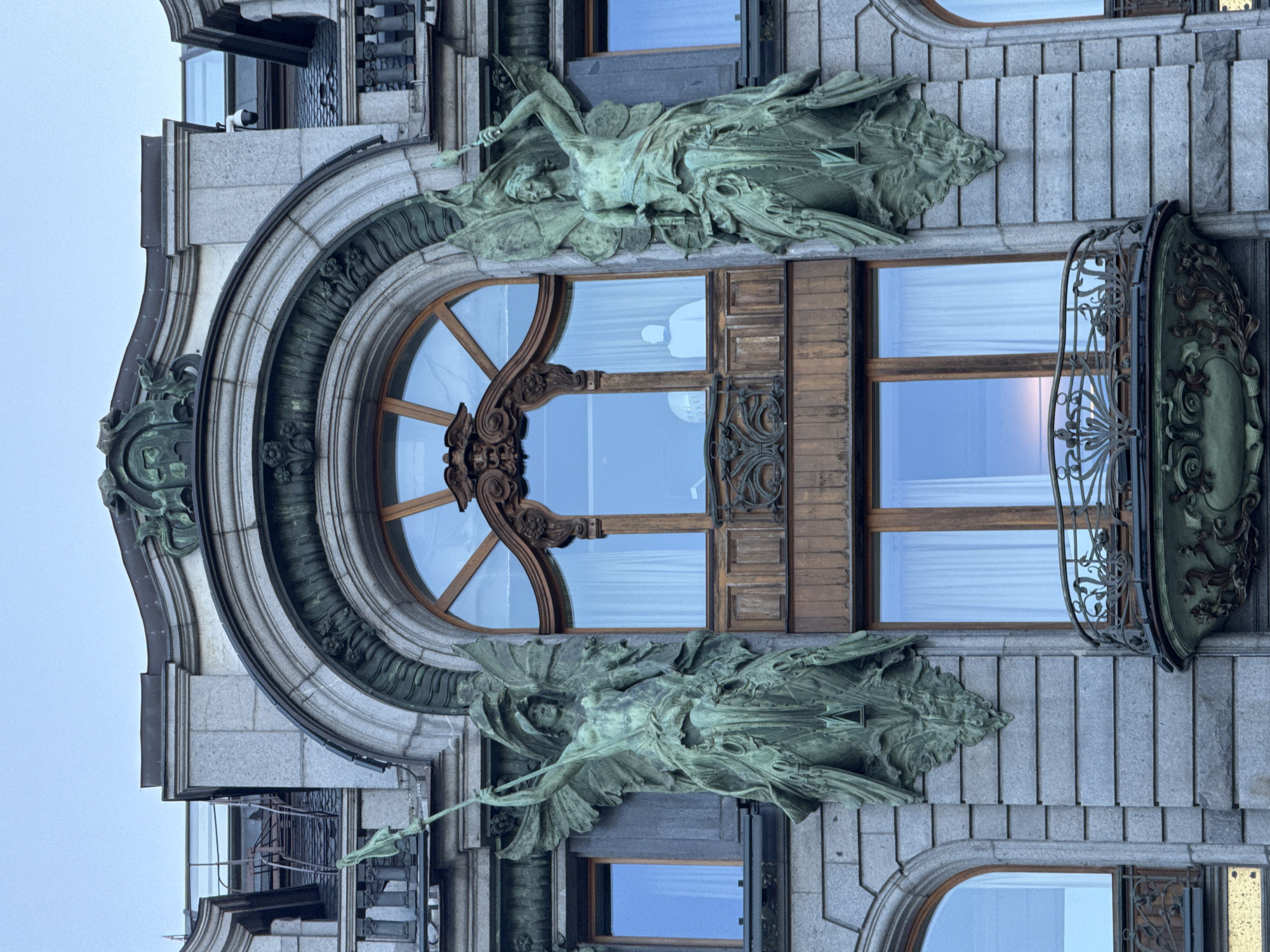 The image shows a detail of the facade of the Singer House, also known as the Dom Knigi, located on Nevsky Prospekt in Saint Petersburg, Russia. The photo focuses on a large, arched window with ornate wooden framing and two green patinated bronze sculptures of winged figures holding staffs, flanking the window. The figures are positioned on either side of the window, contributing to the building's distinctive Art Nouveau style. Below the main window is a smaller balcony with decorative metalwork. The building's stonework is visible, along with other windows and architectural elements. The sky is overcast, suggesting either early morning or late evening.