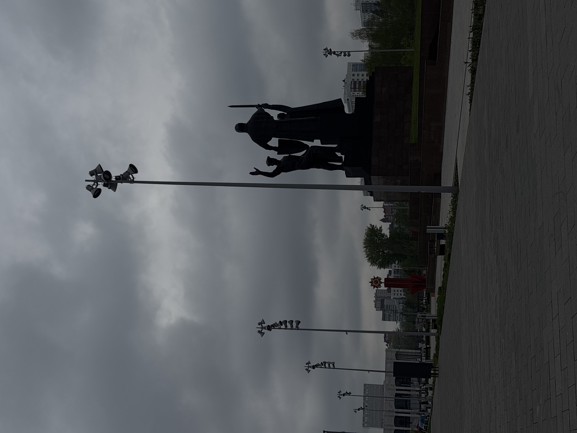 The image depicts a memorial complex with statues in Perm, Russia, against a backdrop of a cloudy sky and urban landscape. The statues are mounted on a dark stone base and represent figures of soldiers or historical figures. Several poles with multiple loudspeakers are visible, as well as a red monument adorned with a Soviet star. The ground is paved with stone blocks, and the photograph appears to have been taken at ground level.