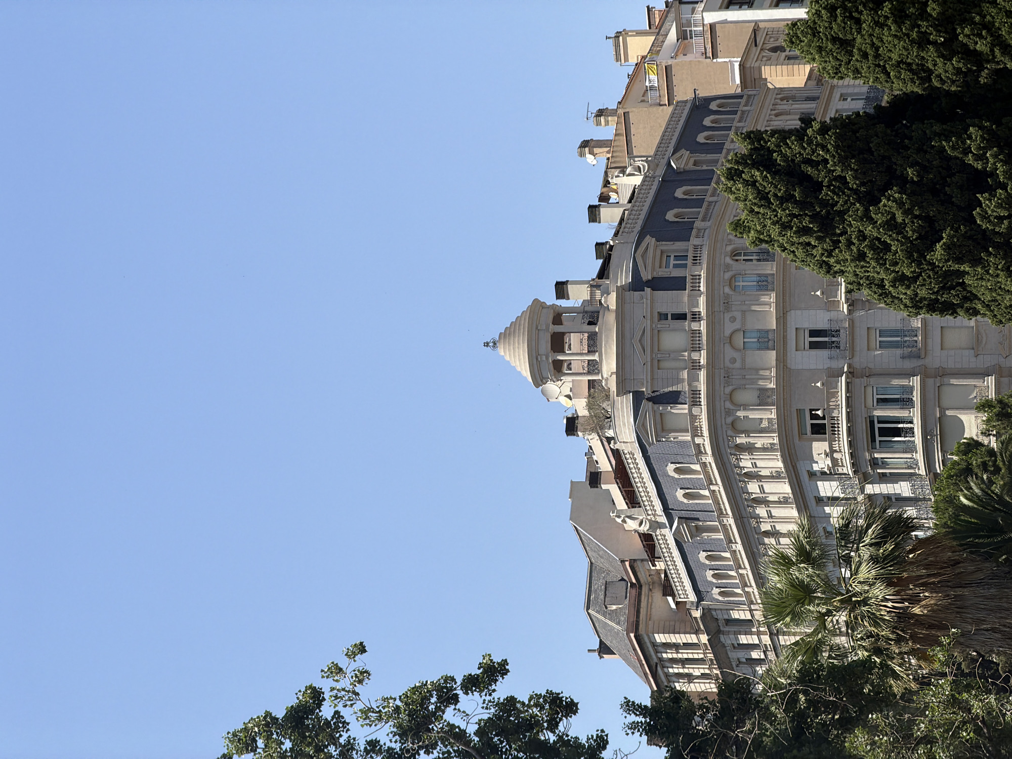 This image captures a section of a curved, ornate building with a light-colored facade against a clear, bright blue sky. The architecture is detailed, featuring multiple windows, balconies with decorative railings, and a domed structure topped with a small spire. The building appears to be several stories high, with varied rooflines and architectural styles visible on different sections. Lush green trees, including what appears to be a palm tree and a dense evergreen, partially obscure the lower and right portions of the building, suggesting a sunlit outdoor setting. The lighting indicates it is daytime, likely with abundant sunshine, casting shadows on the building's intricate details. The image is oriented vertically, giving a sense of looking up at the building. There are no people visible, nor any specific events taking place. The location is identified as Barcelona, Spain, which aligns with the architectural style and the presence of palm trees.