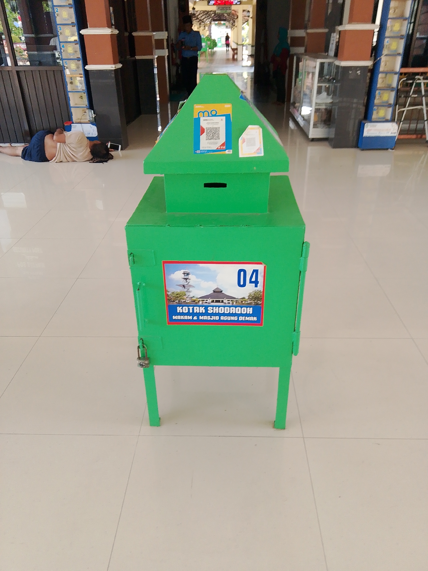 The image shows a green donation box in the foreground, placed on a tiled floor. The donation box has a peaked roof and a slot on the front. A sticker on the box features the number "04" and text that appears to be "KOTAK SHODAQOH MAKAM & MASJID AGUNG DEMAK". In the background, a person is lying on the floor. Further in the background, there are pillars, display cases, and other people. The setting appears to be an indoor public space, possibly a market or a religious site, given the text on the donation box which mentions "Makam & Masjid Agung Demak" (Mausoleum & Great Mosque of Demak), indicating the location is Demak, Indonesia.