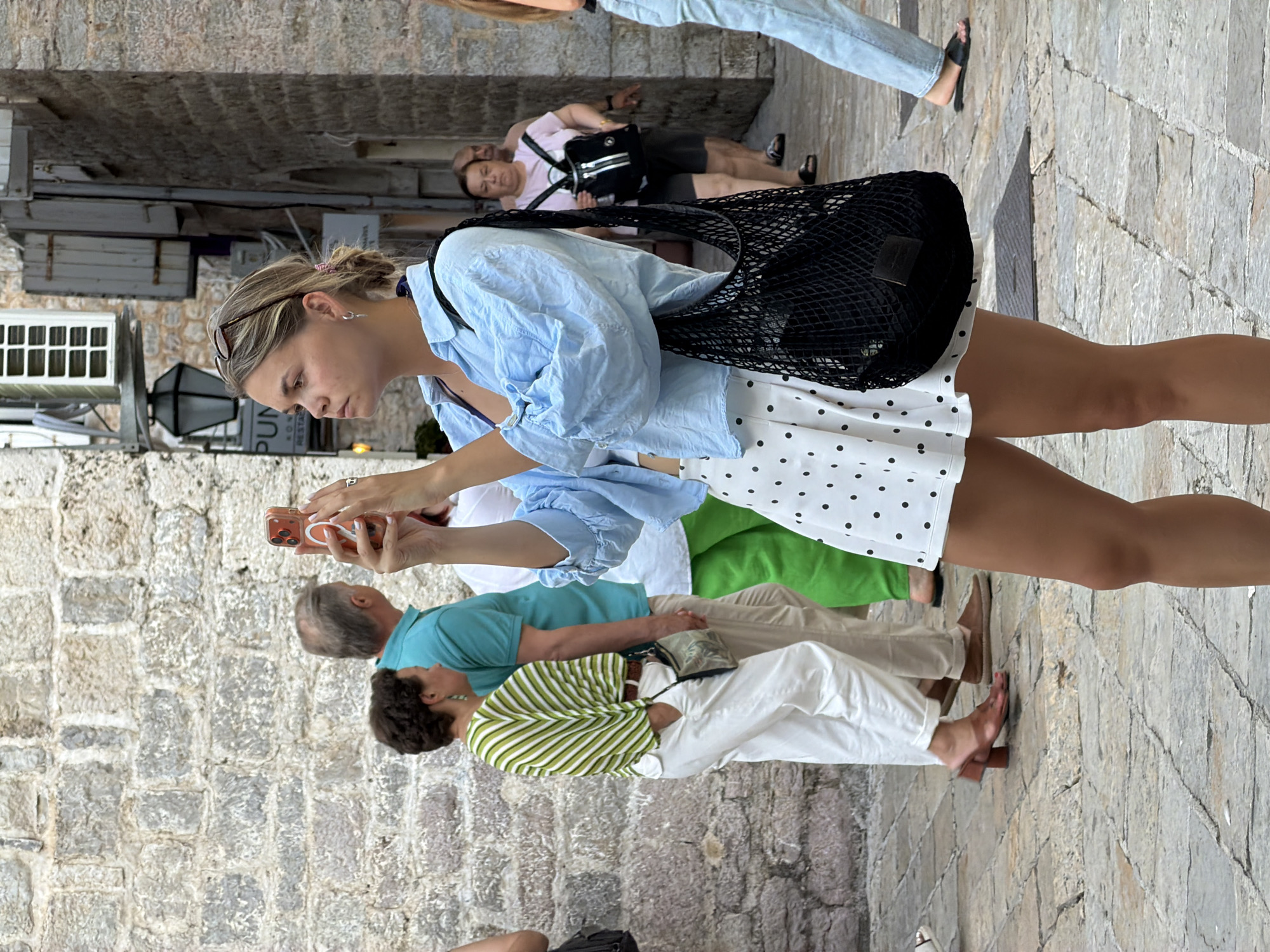 A woman in a blue button-down shirt and a white polka-dot skirt is taking a photo with her phone in an outdoor alleyway in the city of Budva, Montenegro. She is holding a black mesh bag on her right shoulder. In the background, other people are walking, including a man in a turquoise shirt and a woman in a striped green shirt. The alleyway is paved with cobblestones and has stone walls on either side, with some buildings and windows visible. The lighting suggests it is daytime.