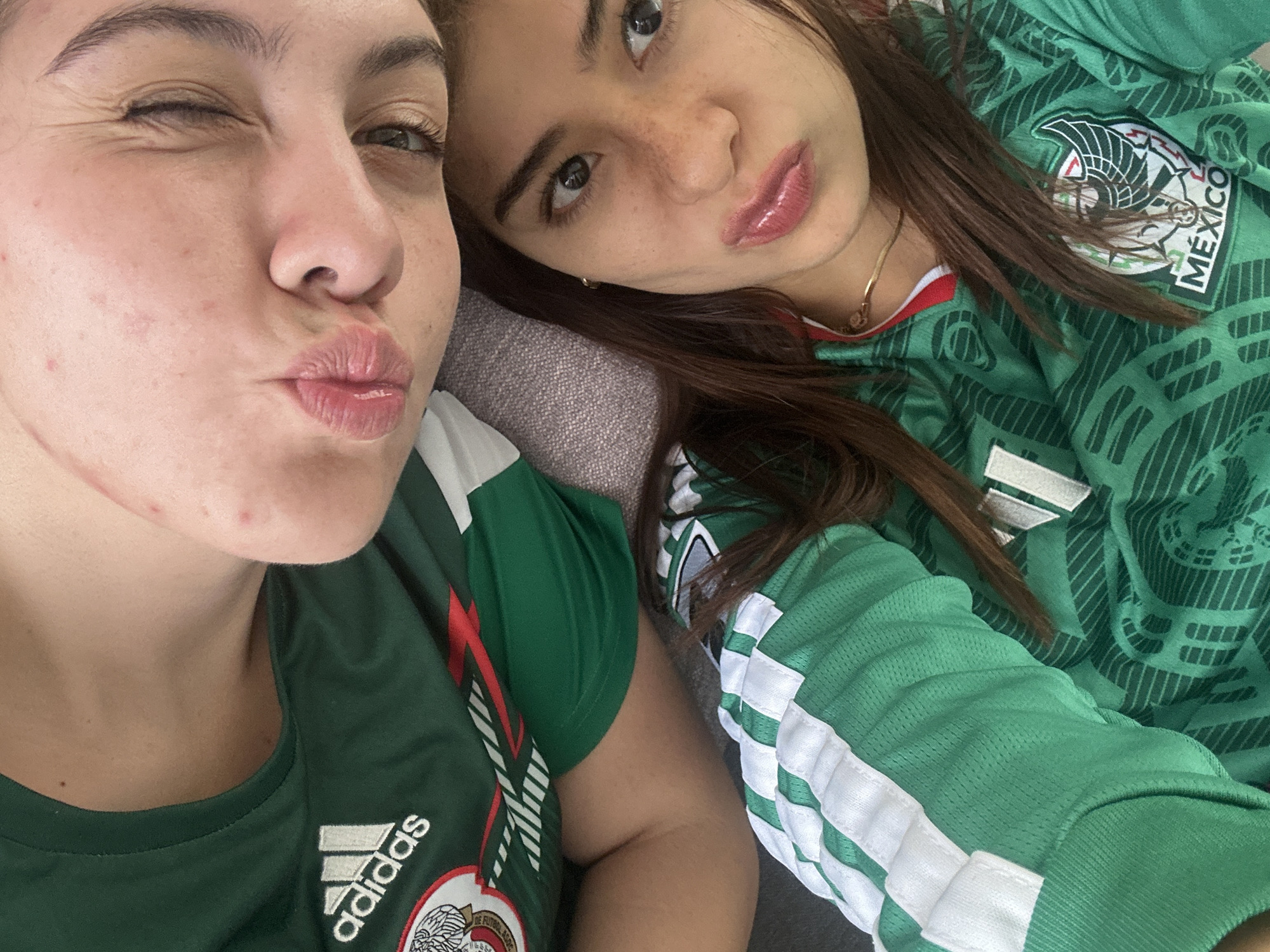 The image is a close-up, high-angle selfie taken by two individuals wearing green Mexican soccer jerseys. One person, positioned higher in the frame, is looking downwards with their eyes closed and lips pursed, appearing to blow a kiss. The other person, positioned lower, is looking directly at the camera with their eyes open and lips also pursed, similar to the first person's pose. Both individuals have clear skin with some blemishes visible on the upper person's face. The jerseys are identical, featuring the Mexican national team crest and Adidas branding. The background appears to be a textured gray surface, possibly a couch or sofa. There is no identifiable information within the image that can be used to determine the location in the city of San Bernardino Tlaxcalancingo, Mexico.