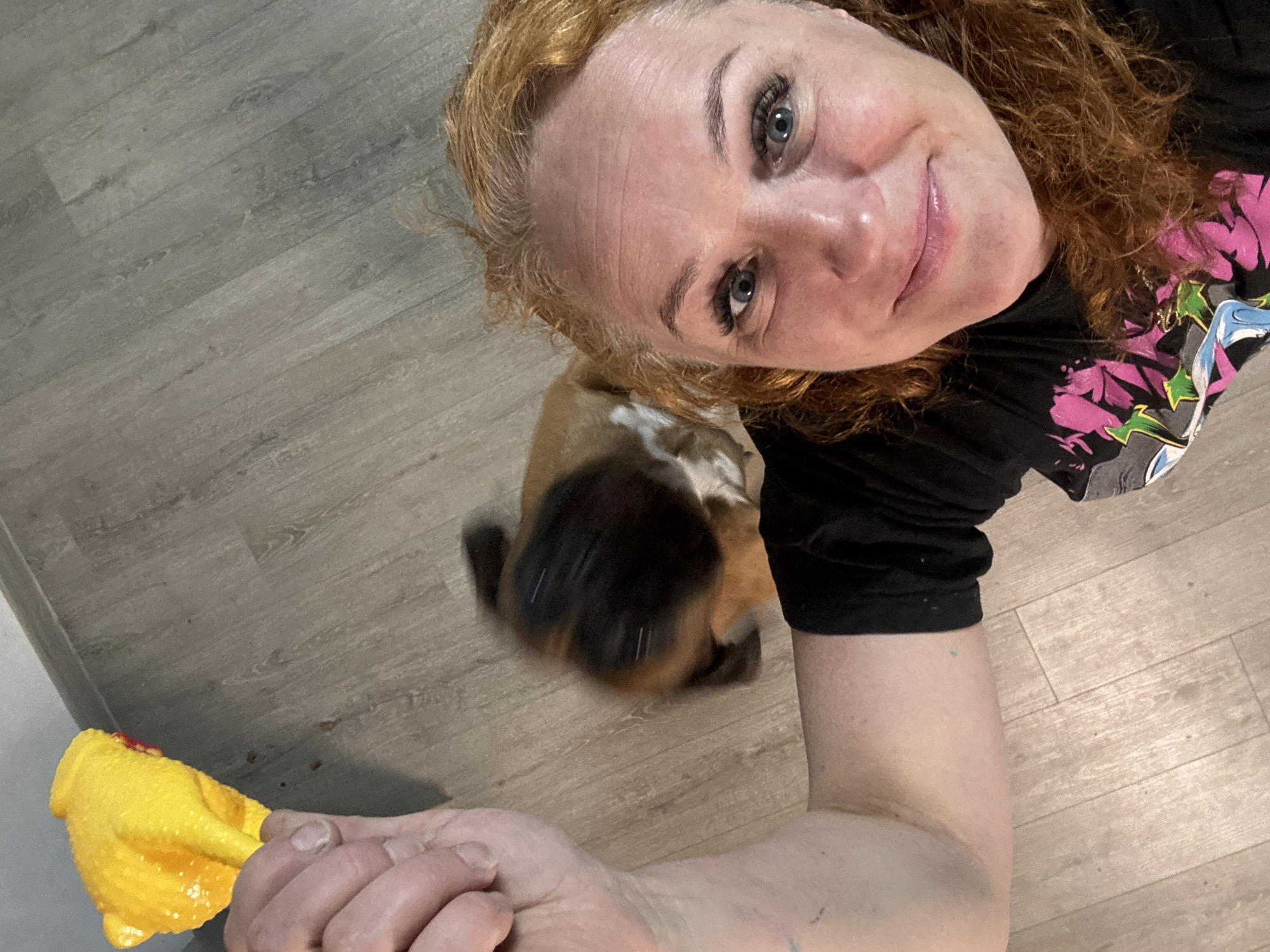 A woman with auburn curly hair is taking a selfie indoors. She is wearing a black t-shirt with a colorful graphic. Her eyes are light blue, and she has fair skin. Below her, a blurred brown and white dog is in motion on a gray wood-look floor. To the left, a yellow toy is visible in her outstretched hand. The precise location within the city of Chaikovsky, Russia, is not ascertainable from the image.