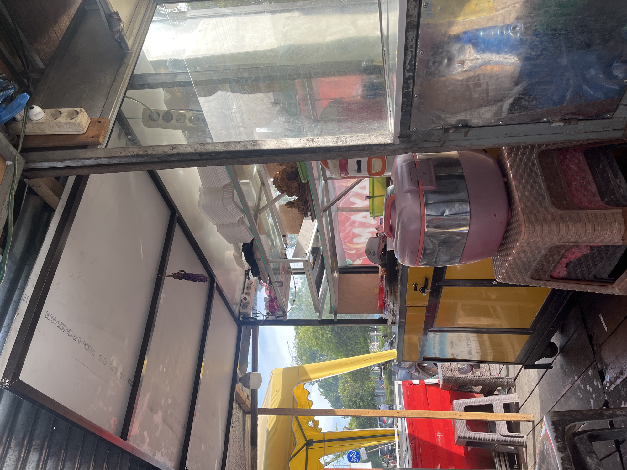 The image depicts an outdoor food stall in Banda Aceh, Indonesia, under a bright sky. The stall is constructed with a metal frame and covered by a yellow awning on the left side and a white panel ceiling on top. A glass-enclosed display area with shelves is visible to the right. The stall is equipped with a food warmer, plastic containers, and what appears to be fried food on a plate. Various seating options, including plastic stools, are present. In the background, trees, vehicles, and a street indicate an urban setting. The overall scene is consistent with a small business operating in an open-air environment.