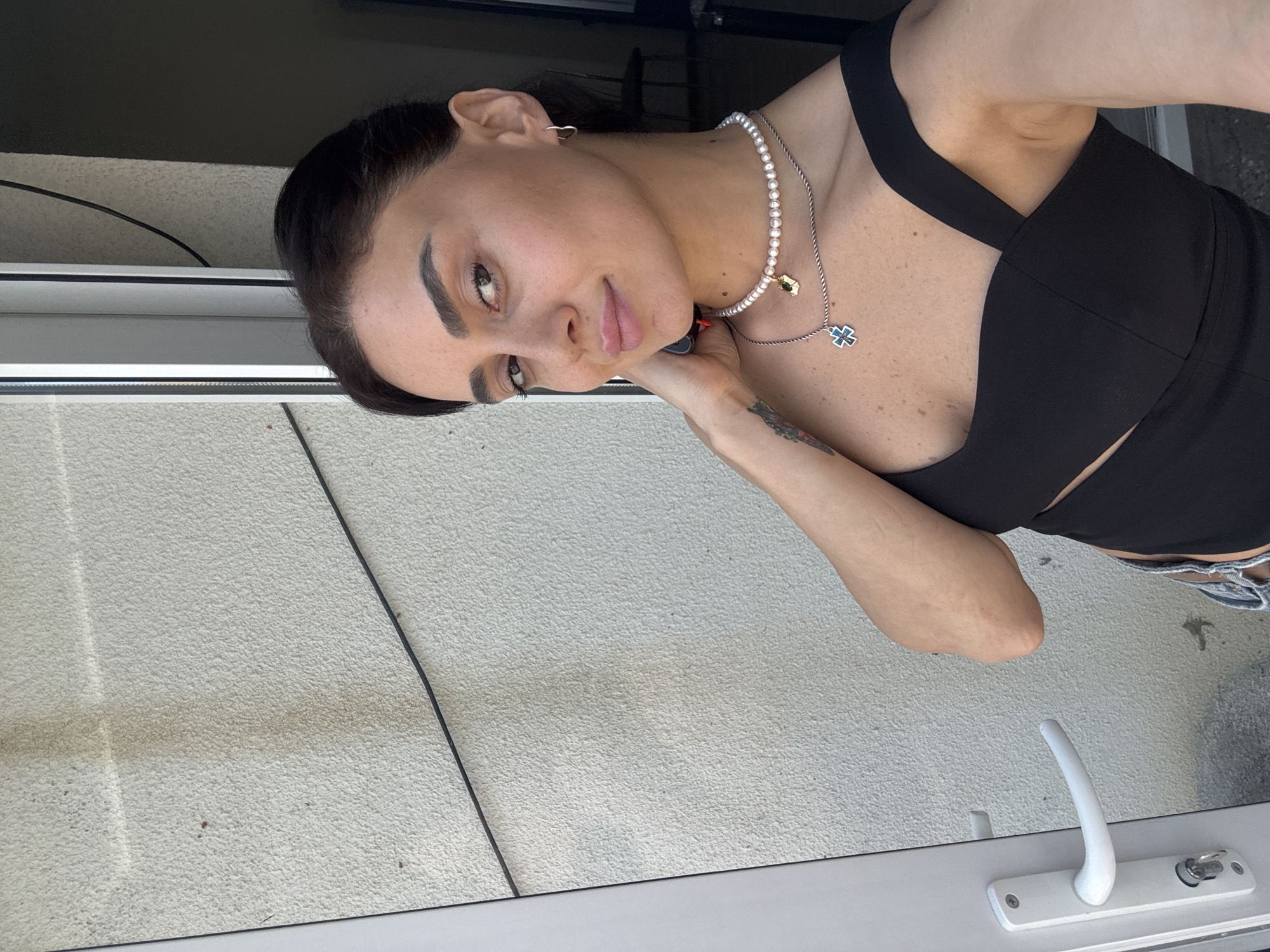The media file shows a woman posing for a photo, with her head tilted and her hand resting on her chin. She has dark hair, styled neatly, and is wearing makeup. She is dressed in a black, sleeveless top and what appears to be denim shorts. A pearl necklace adorns her neck, with a pendant featuring a cross shape. A tattoo is visible on her left forearm.
The background consists of a textured, light-colored wall, possibly stucco or concrete, and a window frame with a white handle and lock mechanism. The lighting suggests it's daytime, possibly outdoors, with natural light illuminating the scene. The overall impression is a casual, personal photograph, likely taken by the subject herself. No text is visible in the image.
Given the location context of Tbilisi, Georgia, the scene could be a balcony, patio, or window area of a residence or establishment in the city. The weather appears to be pleasant, conducive to outdoor photography. The woman's expression is relaxed and engaging, suggesting she is comfortable and enjoying the moment of being photographed.