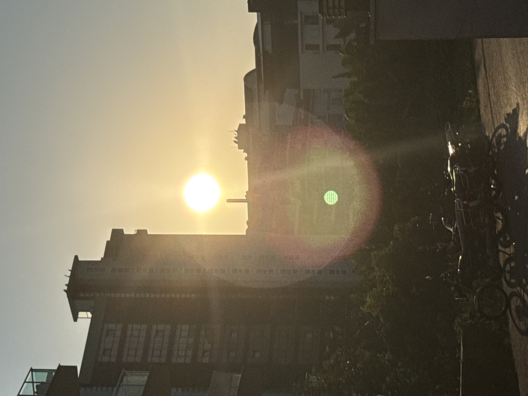 The image shows a street scene in Alanya, Turkey, during sunset. The sun is low in the sky, casting a warm, golden light and creating lens flares. In the foreground, a row of bicycles is parked, their shadows stretching across the pavement. Behind the bicycles, a dark silhouette of trees and foliage can be seen, suggesting a park or garden area. Further back, a tall, modern building with a repeating pattern of windows is illuminated by the setting sun, creating a stark contrast between the light and the building's dark facade. The sky is clear and hazy with the sun's glow. The overall mood is serene and captures the peaceful ambiance of an evening in a city. No people are visible in the frame. There is no visible text in the image.