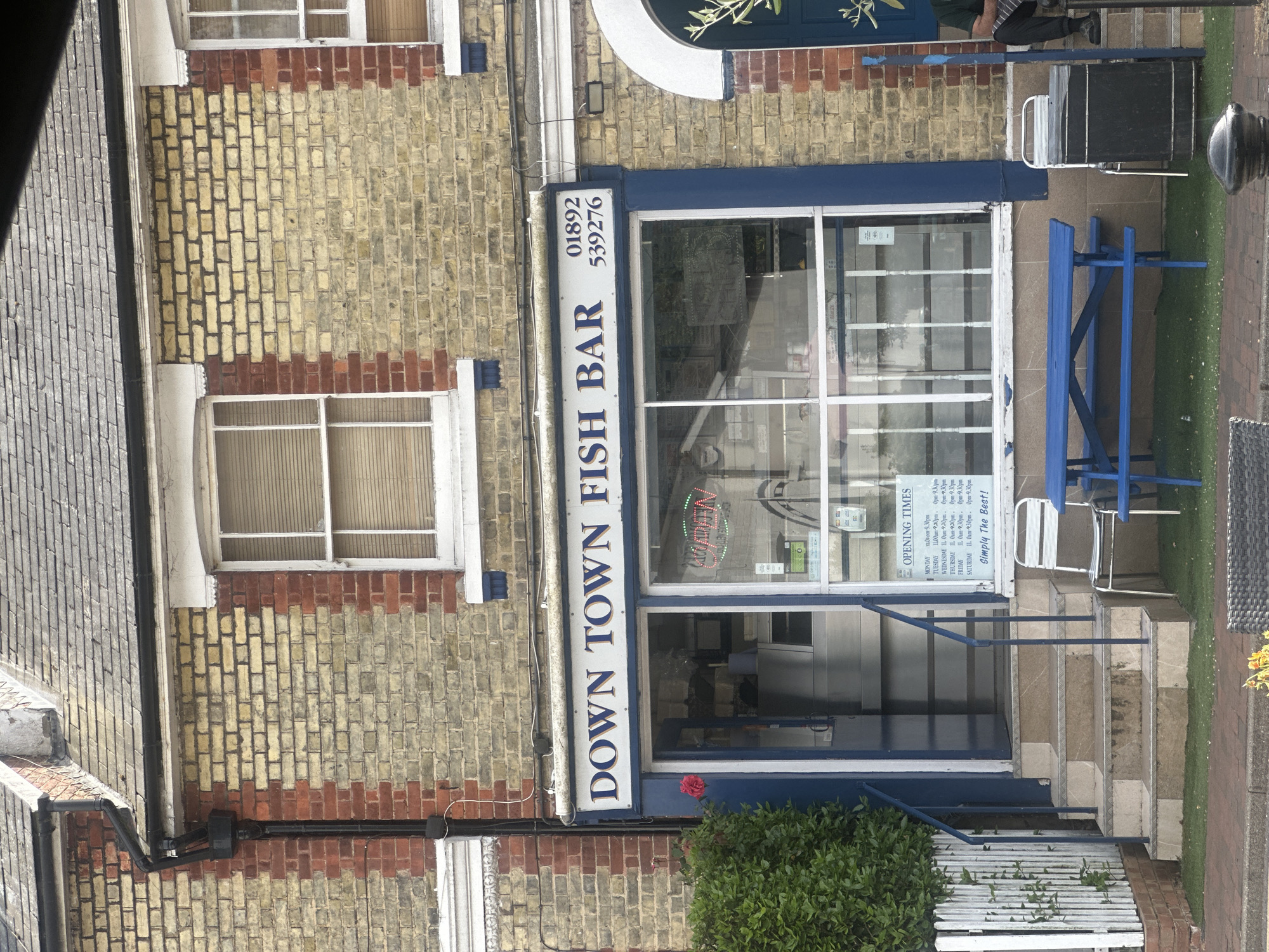 The image depicts the facade of "Down Town Fish Bar," a restaurant located in Tunbridge Wells, United Kingdom. The building features yellow brickwork with red brick accents, and a blue and white sign displaying the business name and telephone number. The front entrance is framed by a blue painted storefront with a large window, revealing the restaurant's interior. Opening times are displayed on a sign. Outside, there is a blue picnic table and chairs, along with a white picket fence and some greenery. A street lamp is visible on the sidewalk in front of the restaurant.