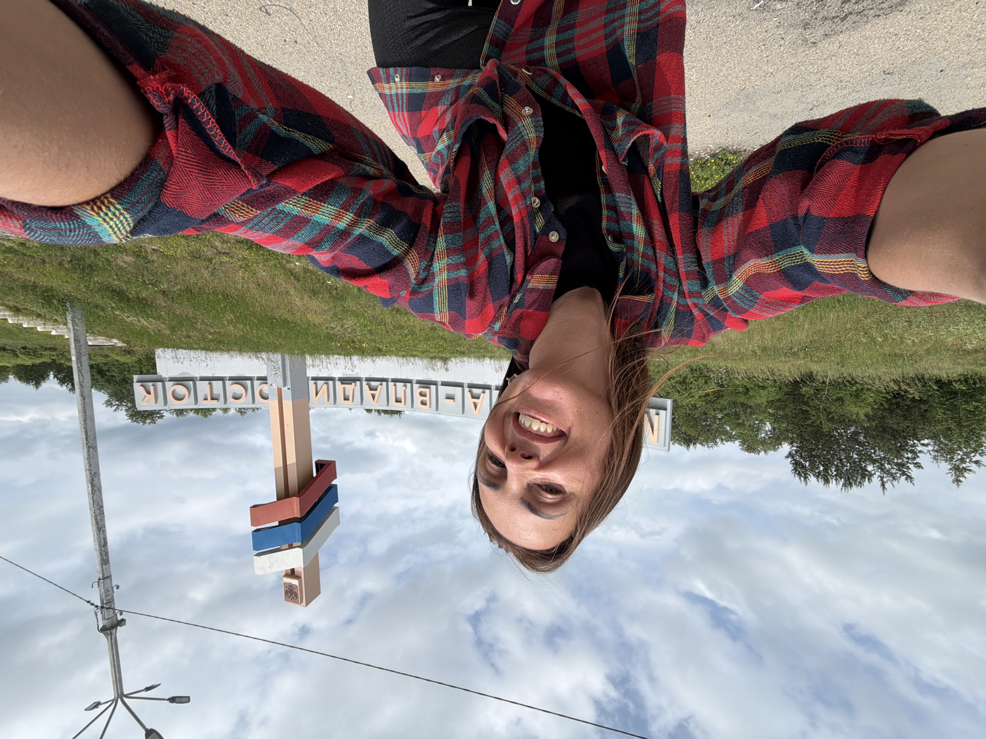 A woman with long brown hair is taking a selfie outdoors. She is wearing a red, blue, and yellow plaid shirt over a black top. Behind her, a sign with Cyrillic lettering reads "МА-ВЛАДИВОСТОК". A pole with three colored stripes (white, blue, and red) stands to the right of the sign, topped by a coat of arms. A lamppost is also visible to the far right. The background consists of a grassy slope, trees, and a cloudy sky. The ground in the foreground is a gravel path.