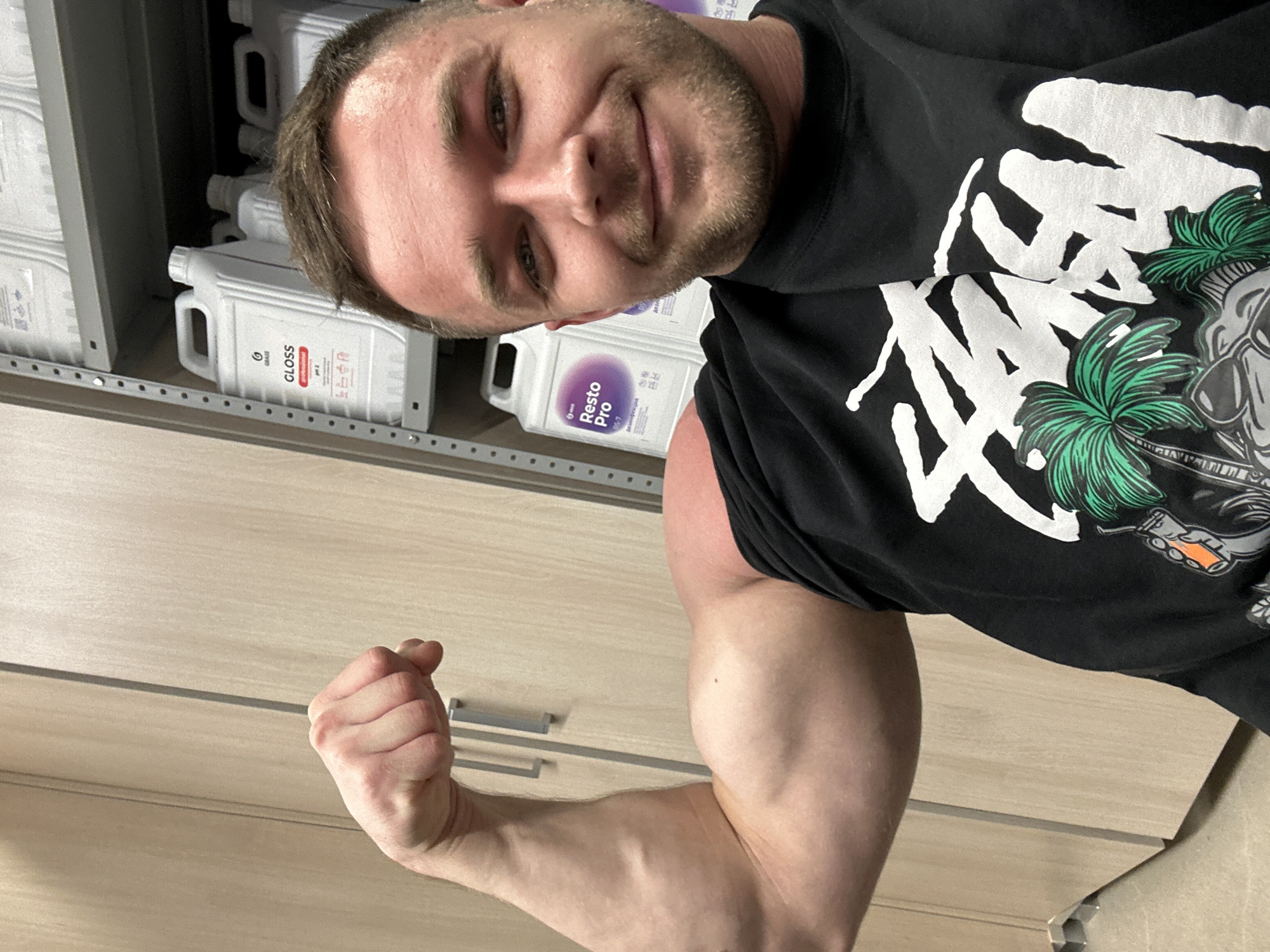 A Caucasian male in a black graphic t-shirt is posing, flexing his right arm and making a fist. Behind him are shelves with several white bottles, labeled "Gloss" and "Resto Pro". A light wood-paneled wall is visible behind his arm. The city location is not discernible from the image.