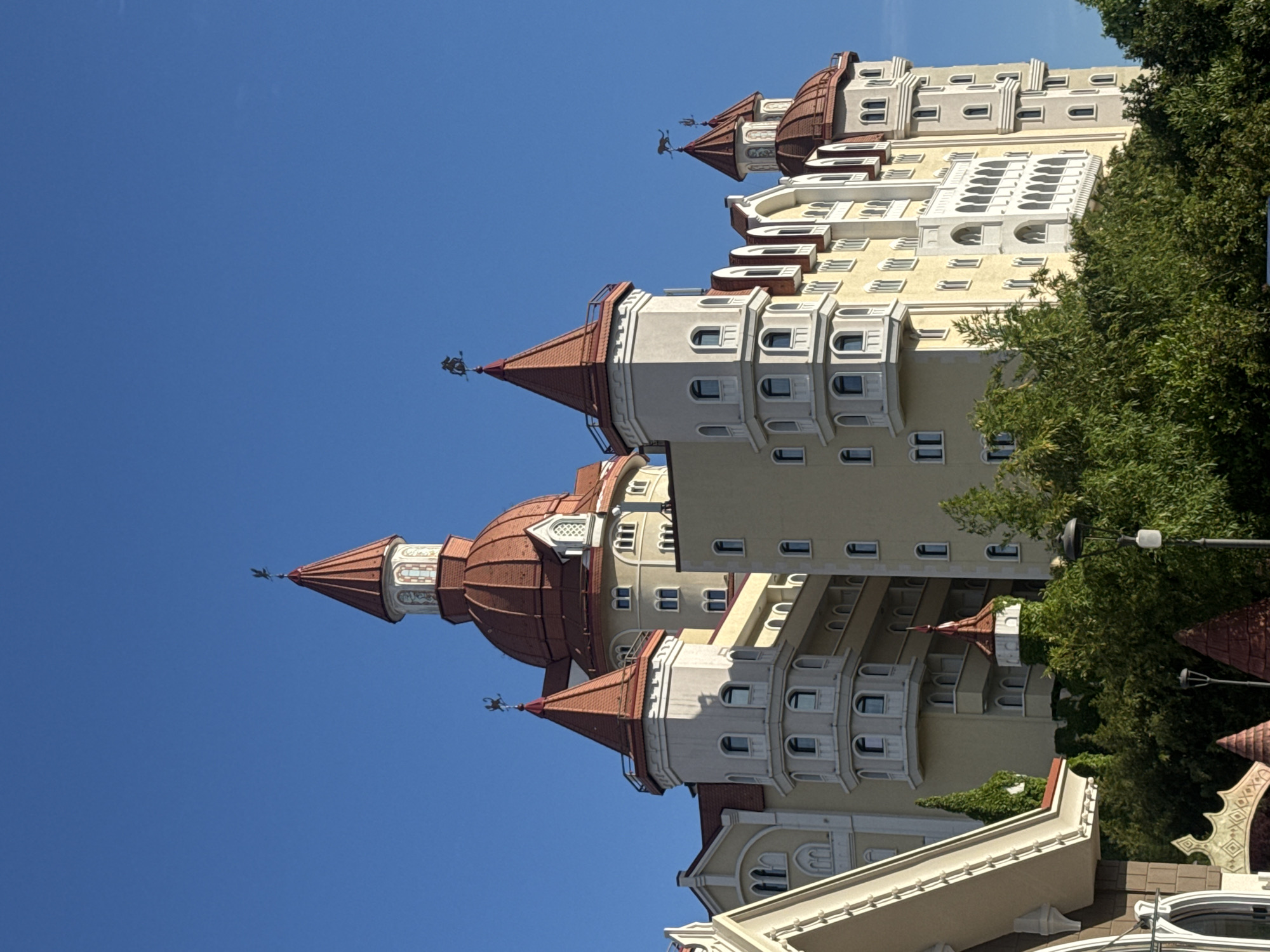 A vertical, eye-level outdoor shot reveals a turreted hotel against a clear blue sky in the city of Sirius, Russia. The hotel's architectural style mimics a medieval castle with multiple towers capped with red-tiled conical roofs. Weather vanes in the shape of mythical creatures adorn the tower peaks. The hotel's facade features a light yellow color scheme, accented by white detailing around the numerous arched windows and decorative moldings. Lush green trees partially obscure the lower right corner of the structure, adding a touch of nature to the scene. The overall ambiance suggests a blend of fantasy and luxury against the backdrop of a clear summer day.