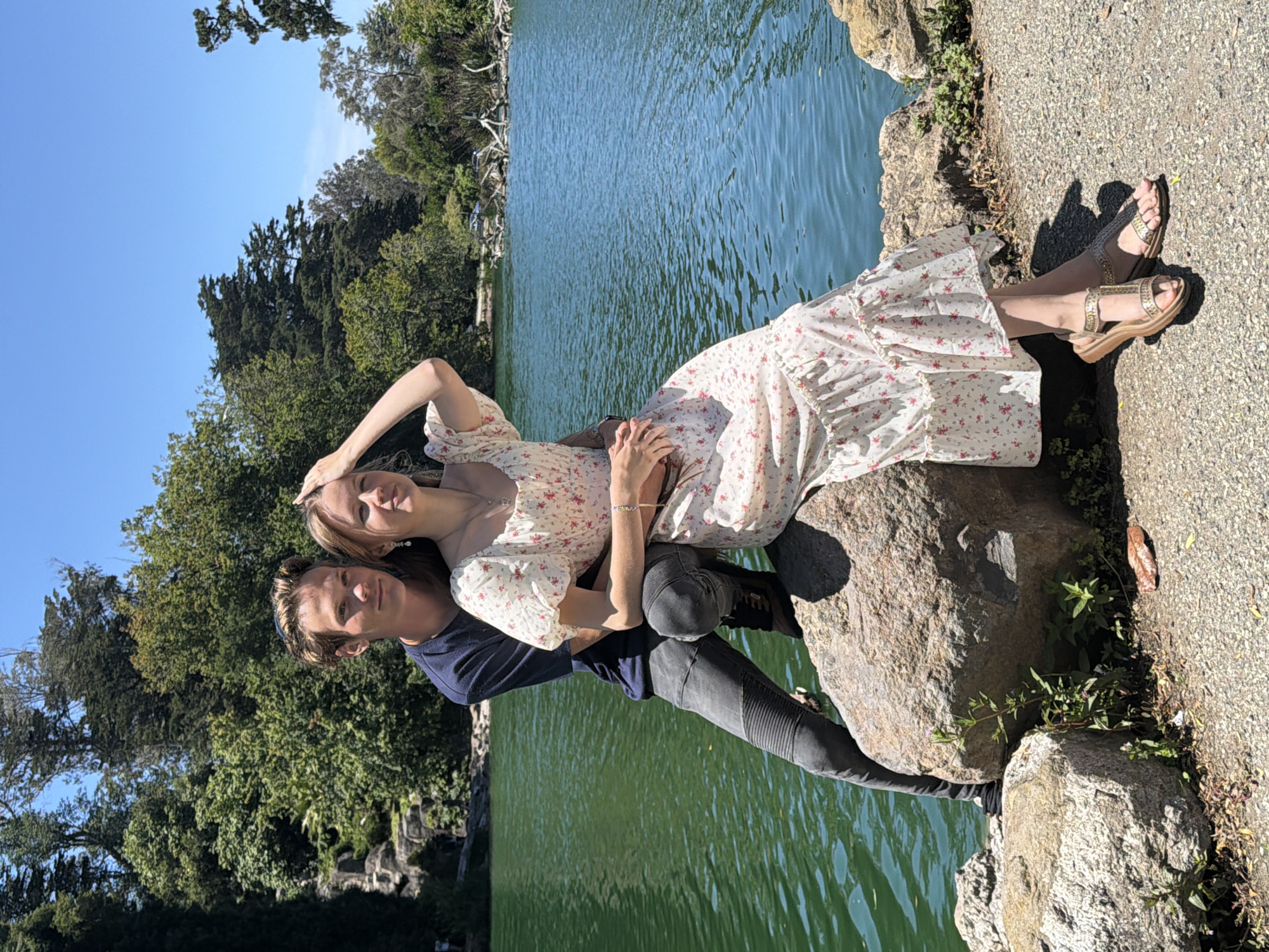 In San Francisco, California, a young couple is pictured on a sunny day by a lake in Golden Gate Park. The man, wearing a navy blue t-shirt and dark grey jeans with a moto-style ribbed detail on the thighs, is seated and has his arm around the woman. The woman, in a white floral dress with short, puffed sleeves and a ruffled hem, is sitting on a large, rough-hewn rock. She has her hand on her head, her fingers touching her hair. The lake behind them has green, rippling water, and the shoreline is lined with rocks and lush green trees. The sky is a clear, bright blue, indicating good weather.