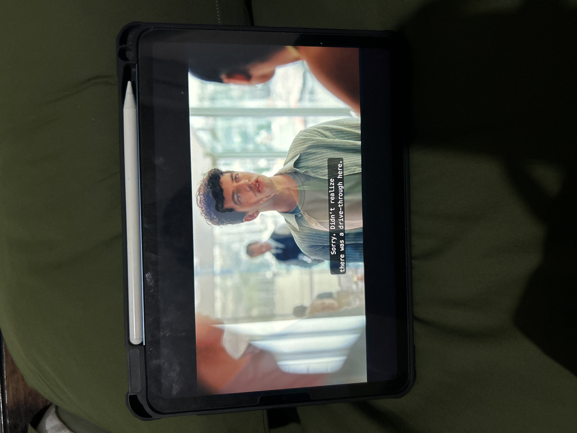 The image shows a tablet device, likely an iPad, resting on a dark green, textured surface, possibly fabric. The tablet is displaying a video scene with subtitles. In the video, a young man with curly brown hair and a light green striped shirt is speaking. He appears to be in an indoor setting with large windows, and there are other figures blurred in the background, including one man in a dark vest and tie. The subtitles read, "Sorry, Didn't realize there was a drive-through here." A white stylus is attached to the top edge of the tablet case. The overall scene is dimly lit, with shadows cast on the green surface, suggesting the room is not brightly illuminated. There is no visible indication of the location being in Sukawati, Indonesia.