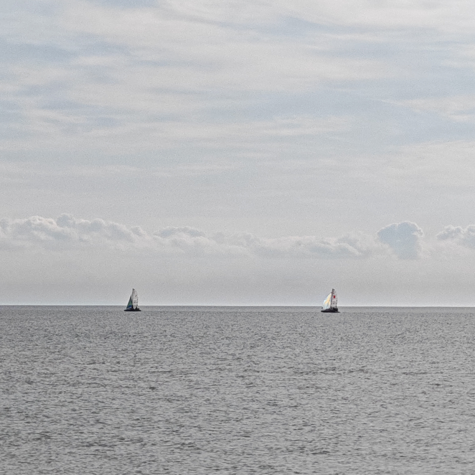 The image shows a seascape with two sailboats on a flat, gray sea under a pale, cloudy sky. The horizon line is roughly in the middle of the frame. One sailboat is on the left, and the other is on the right. The sails of the boats are visible against the sky. The location is in the Мысовское сельское поселение, Ukraine.