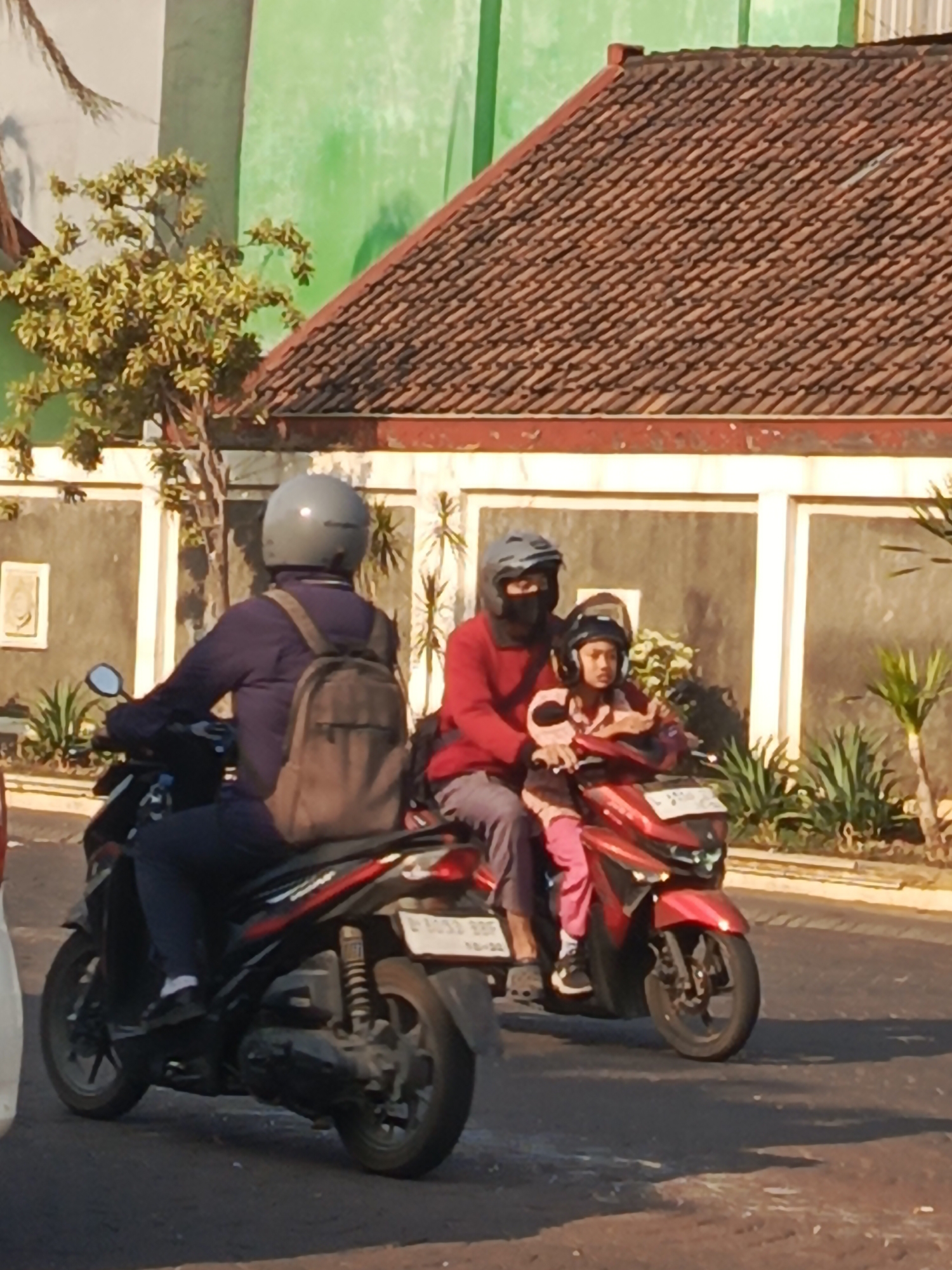 In Boro, Indonesia, three individuals are riding on two motorcycles on an asphalt road. The first motorcycle, to the left, is ridden by an adult wearing a grey helmet, a dark blue jacket, and dark pants, with a tan backpack. The second motorcycle, to the right, carries two individuals. The driver, an adult, wears a dark helmet and a red long-sleeved shirt, and dark pants. A child, wearing a black helmet and a light pink outfit, is seated in front of the driver. The motorcycles are moving from left to right across the frame. The background features a building with a reddish-brown tiled roof and a light-colored wall, with green foliage visible to the left and right. The lighting suggests it is daytime.