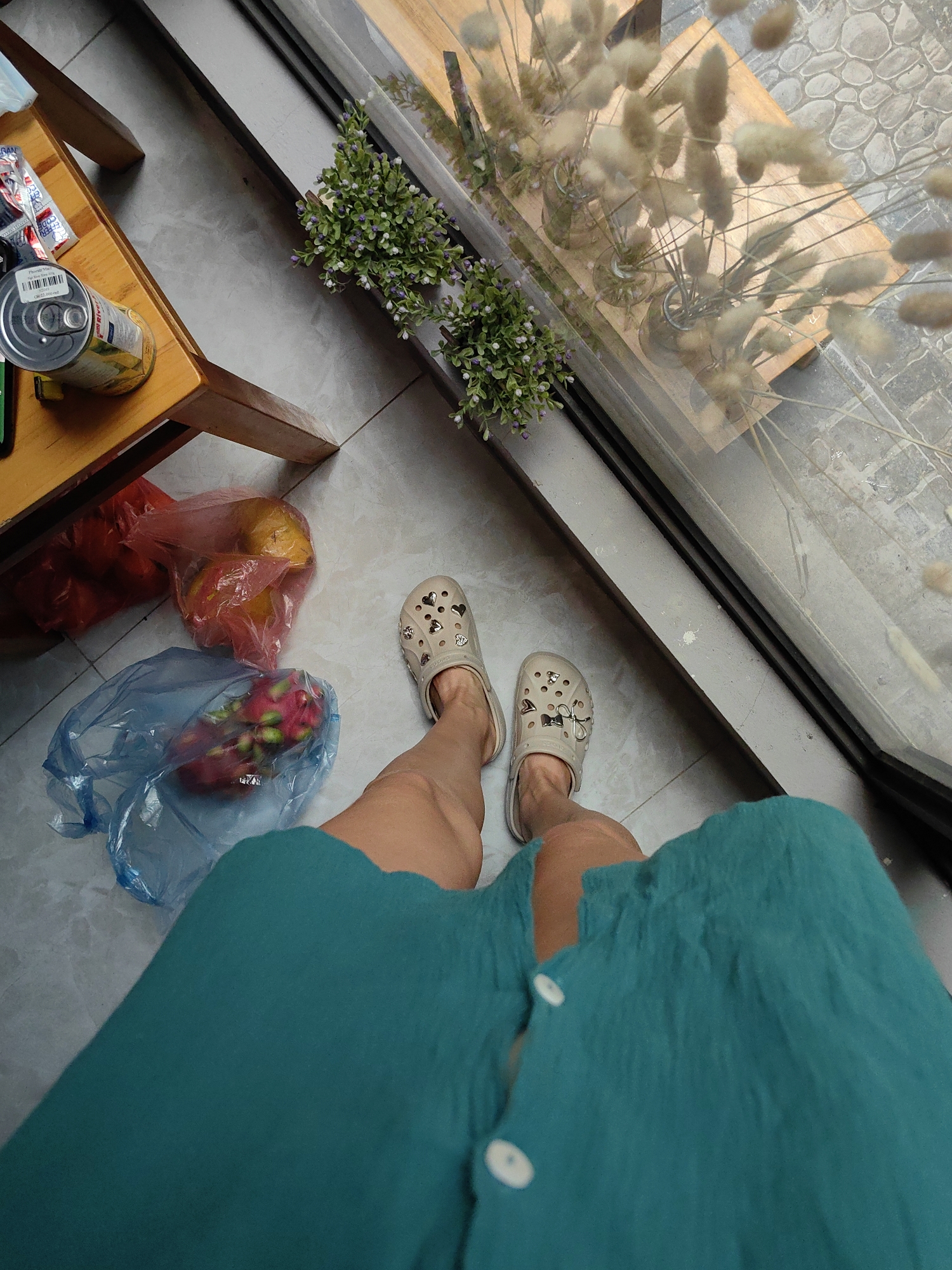 This high-angle, first-person photograph captures a person's lower body, dressed in a teal or turquoise garment and light beige Crocs-style sandals decorated with heart and bow charms. The person stands on a light grey tiled floor next to a large glass window or partition.
To the left, a small wooden table displays a silver can, partially obscured by a barcode, and another yellow container. A small packet, possibly labeled "COFFEE," is also visible. On the floor beneath the table, two plastic bags contain fresh fruit: a red bag holds yellow mangoes, and a blue bag contains red dragon fruit.
Along the window ledge, small potted plants with delicate purple and white flowers are arranged. Through the glass, an outdoor or semi-outdoor area is visible, featuring a wooden table and a stone-paved ground. Several glass vases with dried, fluffy ornamental grasses are also seen on the outdoor table. The scene is brightly lit by natural light, suggesting daytime. The casual attire, fresh fruit, and cafe-like setting blend well with the context of Hà Nội, Vietnam, depicting a relaxed indoor environment with a view to the outside.