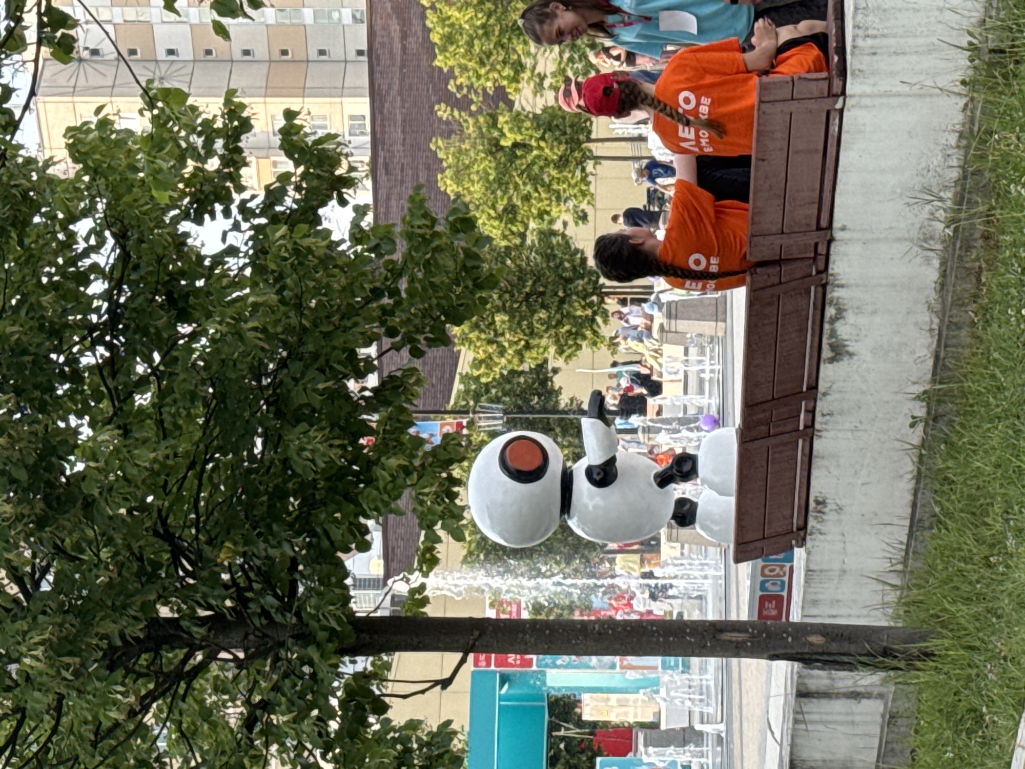 The image depicts an outdoor scene in Zelenograd, Russia, likely a public square or park. In the foreground, a park bench is occupied by three individuals, two of whom are young women wearing orange t-shirts with Cyrillic text on the back. The third person, partially obscured, is also wearing an orange t-shirt. To the left of the bench, a small, white robot statue with a red circular eye stands near a fountain with several water jets. The background features lush green trees, a modern building with a grid-like facade, and more fountains. The overall atmosphere suggests a pleasant, possibly event-filled day in a city environment.