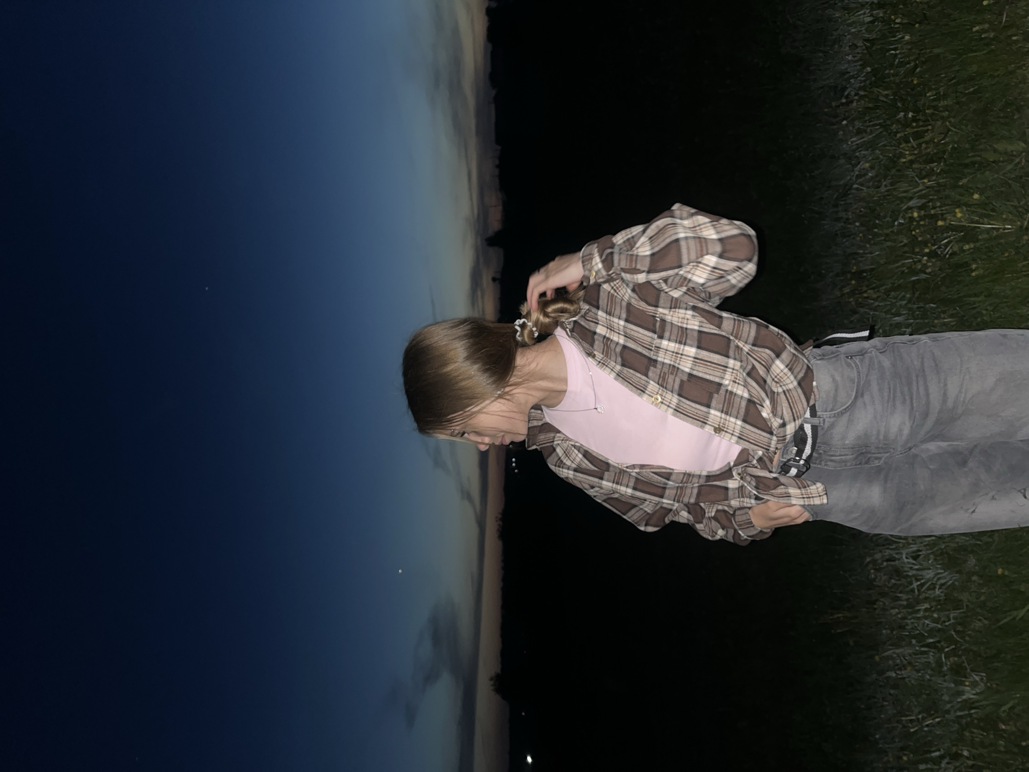 A young woman stands outdoors at dusk or night. She is wearing a pink t-shirt, a brown and white plaid shirt, and gray jeans. She is pulling her hair back into a ponytail. The background shows a dark blue sky on the left and a dark green area on the right, likely grass. The location cannot be determined as Фоки, Russia from the image.
