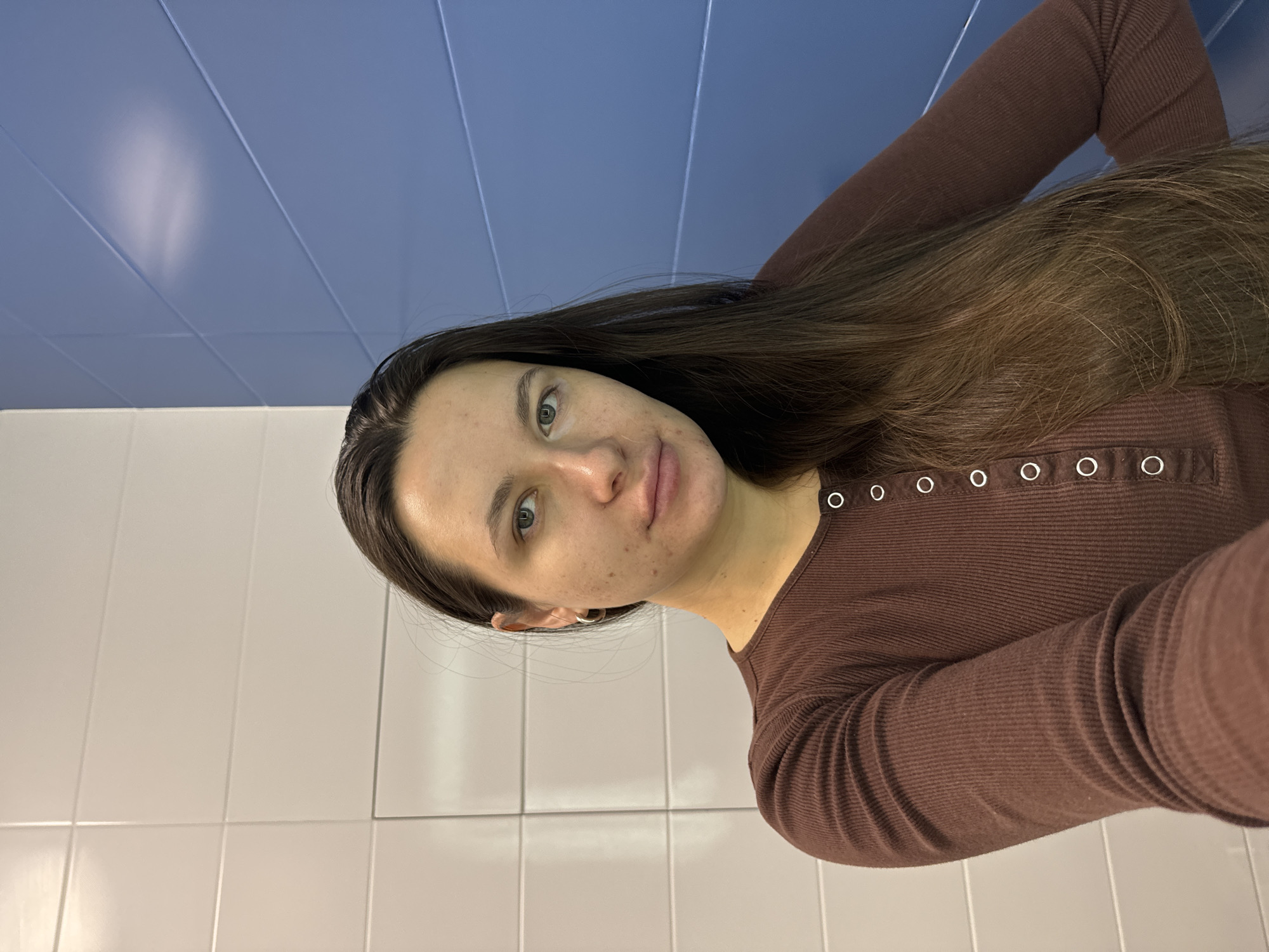 A person is shown in a selfie. The background consists of white tiled walls and a blue ceiling with paneling. The person has long, brown hair and is wearing a brown, long-sleeved shirt with button detailing at the neckline. The person is looking directly at the camera. This image was taken in a bathroom. There is no information that allows for the determination of the specific location in the city of Odintsovo, Russia.