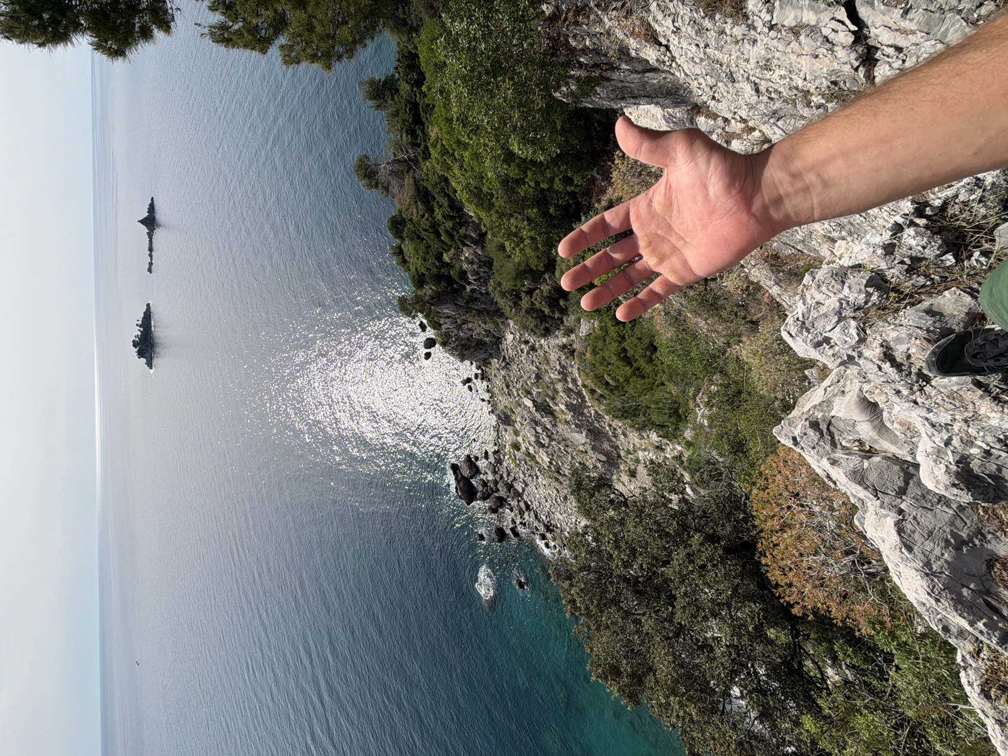 A person stands on a rocky cliff overlooking the Adriatic Sea near Petrovac centar, Montenegro. The person's right hand extends outward. The coastline is rocky and covered with green vegetation. Two small, rocky islands are visible in the distance. The water is calm with sunlight reflecting off the surface.