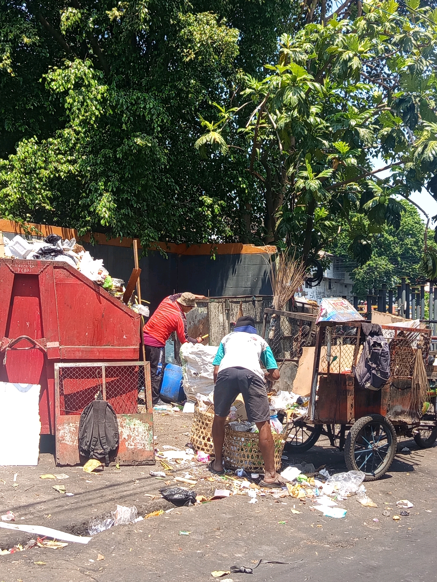 In Semarang, Indonesia, two individuals are engaged in waste management near a large, red dumpster overflowing with refuse. The scene is set on a street littered with garbage, with a dense canopy of green trees overhead.
The first individual, wearing a red long-sleeved shirt and dark pants, is partially visible, interacting with the contents of the dumpster. They appear to be reaching into the dumpster or sorting through waste.
The second individual, facing away from the camera, is dressed in a white t-shirt with teal accents and dark shorts. They are crouched down, sorting through a large woven basket filled with trash that is situated on the ground. This basket is next to a rusty, wooden cart with two large spoked wheels, which is also laden with various items and appears to be a mobile collection point for waste. A black backpack is hanging on a metal grate structure to the left of the red dumpster.
The environment suggests an outdoor, possibly informal, waste collection or sorting area. The ground is asphalt, covered with a significant amount of scattered debris. Buildings and more trees are visible in the background, indicating an urban or semi-urban setting. The lighting suggests it is daytime, with sunlight filtering through the trees.